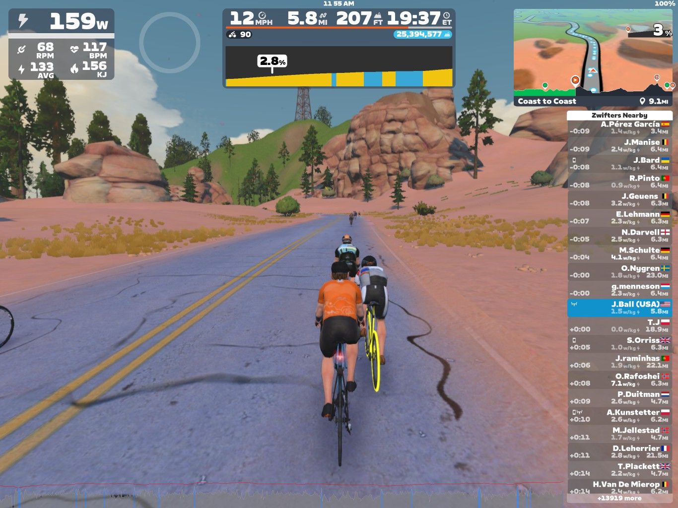 A digital cycling scene shows a group of cyclists riding on a road surrounded by rocky and hilly terrain. The cyclists are in a virtual environment, with stats displayed on the screen, including speed, distance, and power output. The landscape features pink earth and scattered trees.