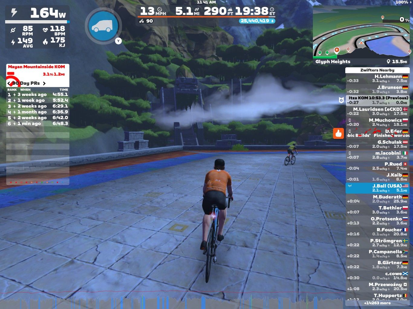 A cyclist wearing an orange jersey rides on a virtual road in a mountainous landscape, with a scoreboard displaying performance stats, nearby cyclists, and recent achievements. The scene includes lush greenery and a water feature in the background.
