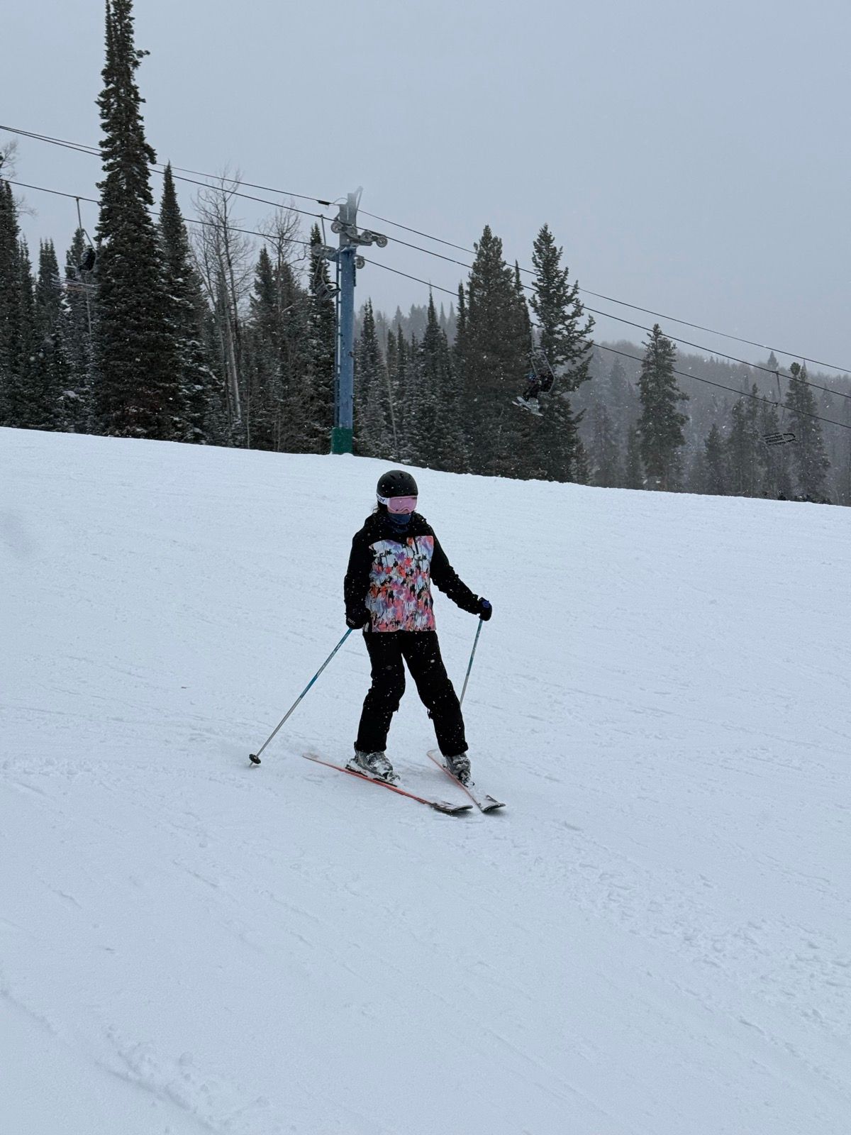 A person skiing on a snowy slope in Logan, UT. The skier is wearing a colorful jacket and black pants, with a helmet and mask. Snow-covered trees and a ski lift are visible in the background.