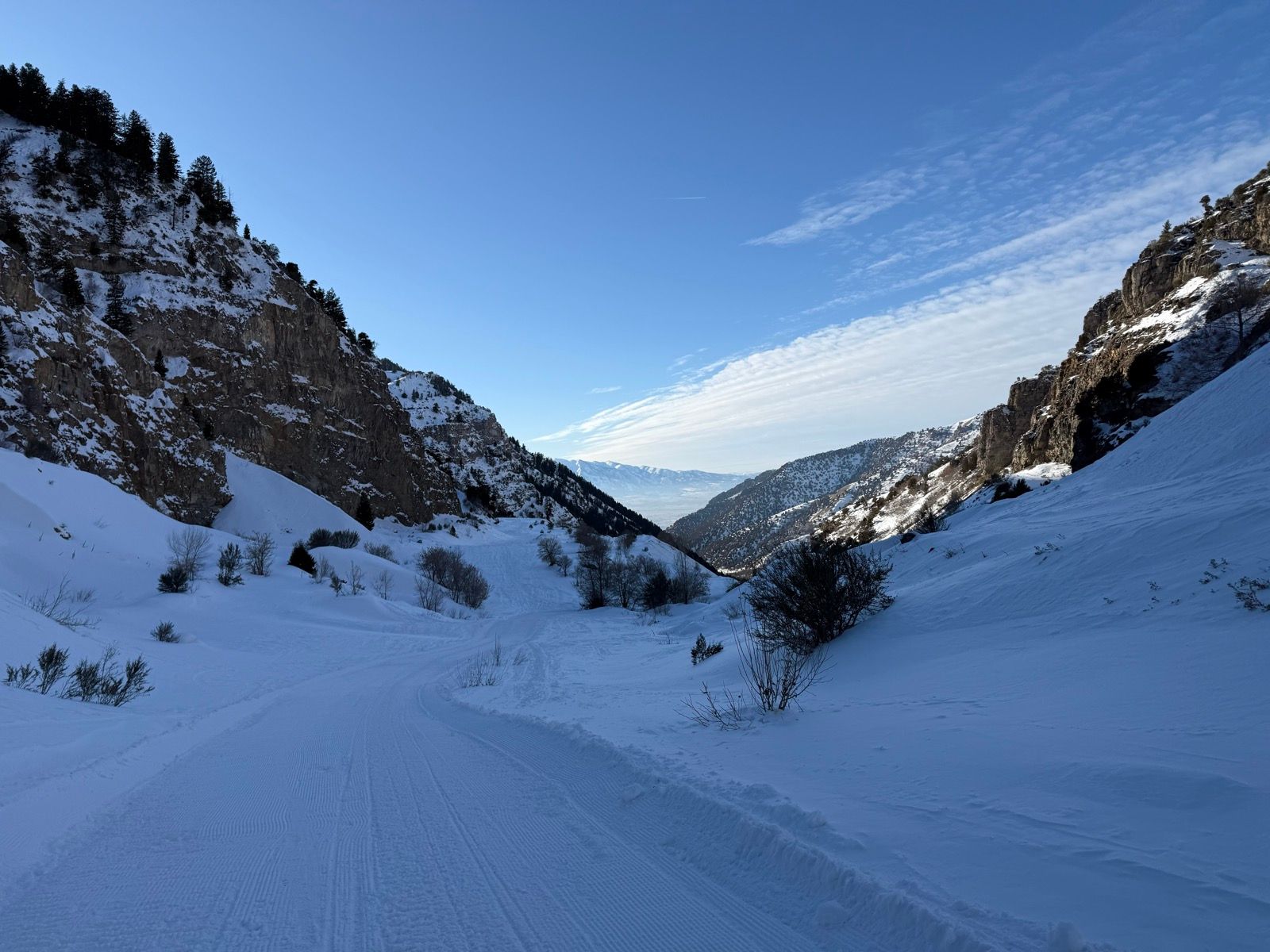 A snow-covered road winds through a canyon surrounded by steep, rocky slopes and scattered trees under a clear blue sky in Providence Canyon, Utah.