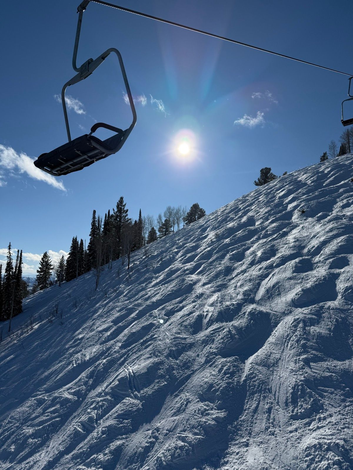 A ski lift chair is suspended above a snowy slope under a bright sun. The hillside features undulating snow patterns and scattered trees in the background, captured against a clear blue sky.