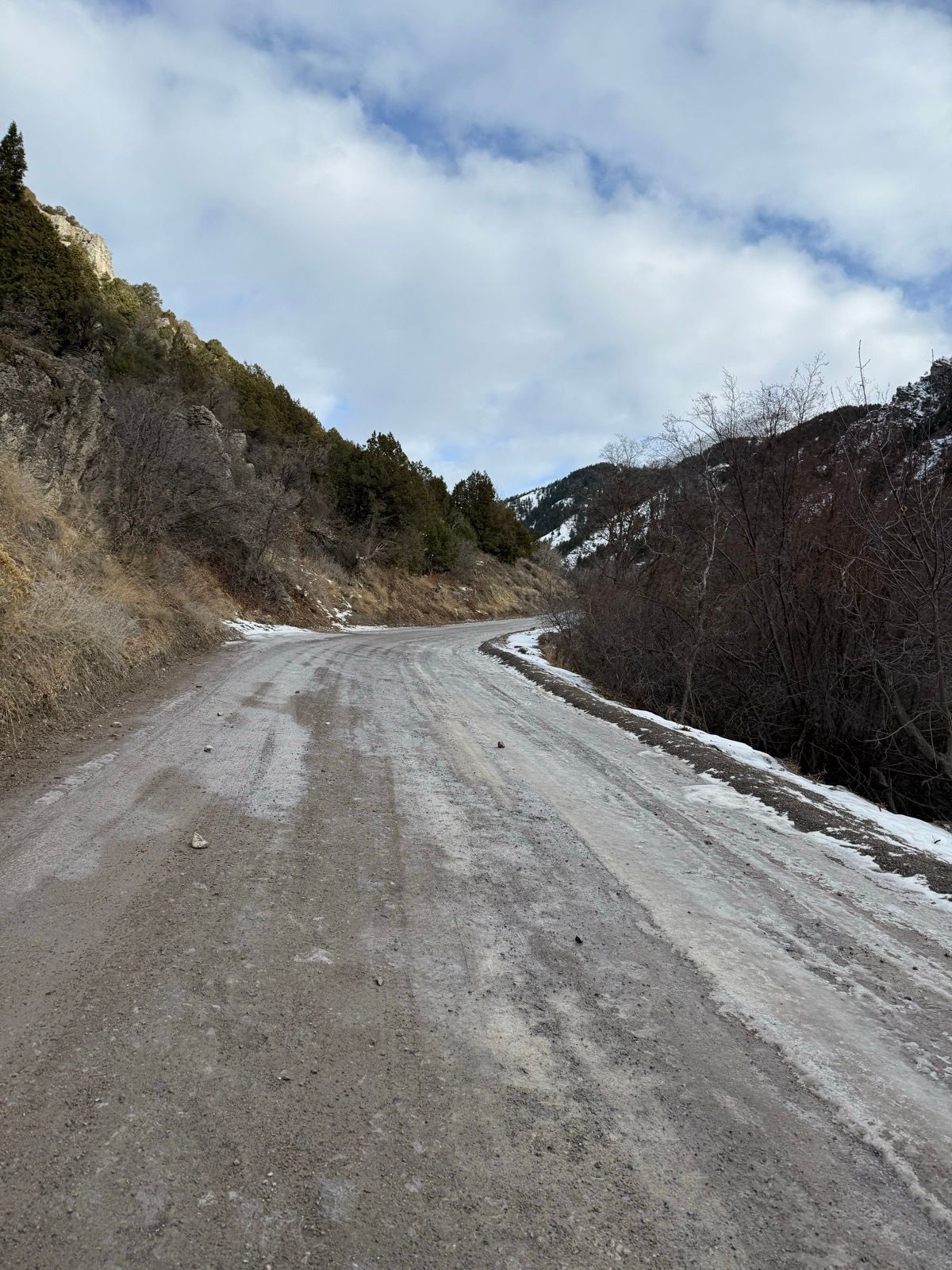 A winding dirt road surrounded by rocky terrain and sparse vegetation, with patches of snow visible on the ground. The scene is set under a cloudy sky, showcasing the mountainous landscape of Logan, Utah.