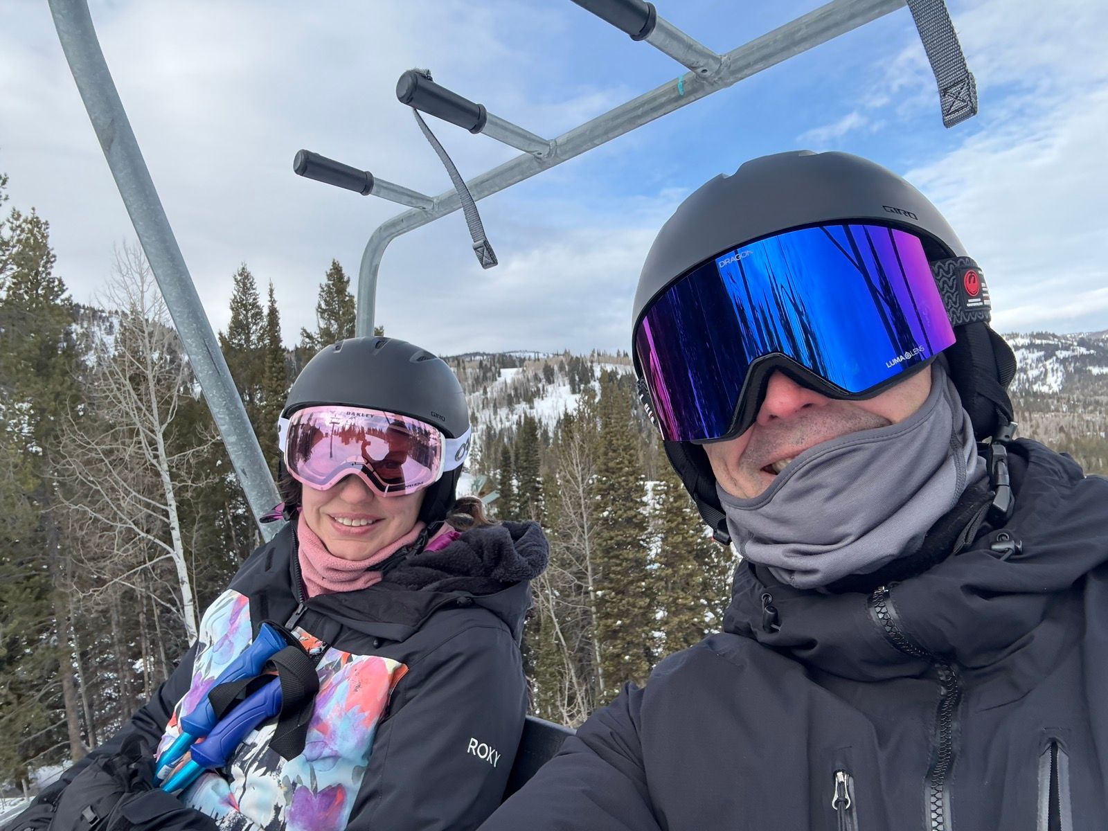 A woman and a man smiling while riding a ski lift. Both are wearing helmets and ski goggles. The background shows snow-covered mountains and evergreen trees under a cloudy sky.