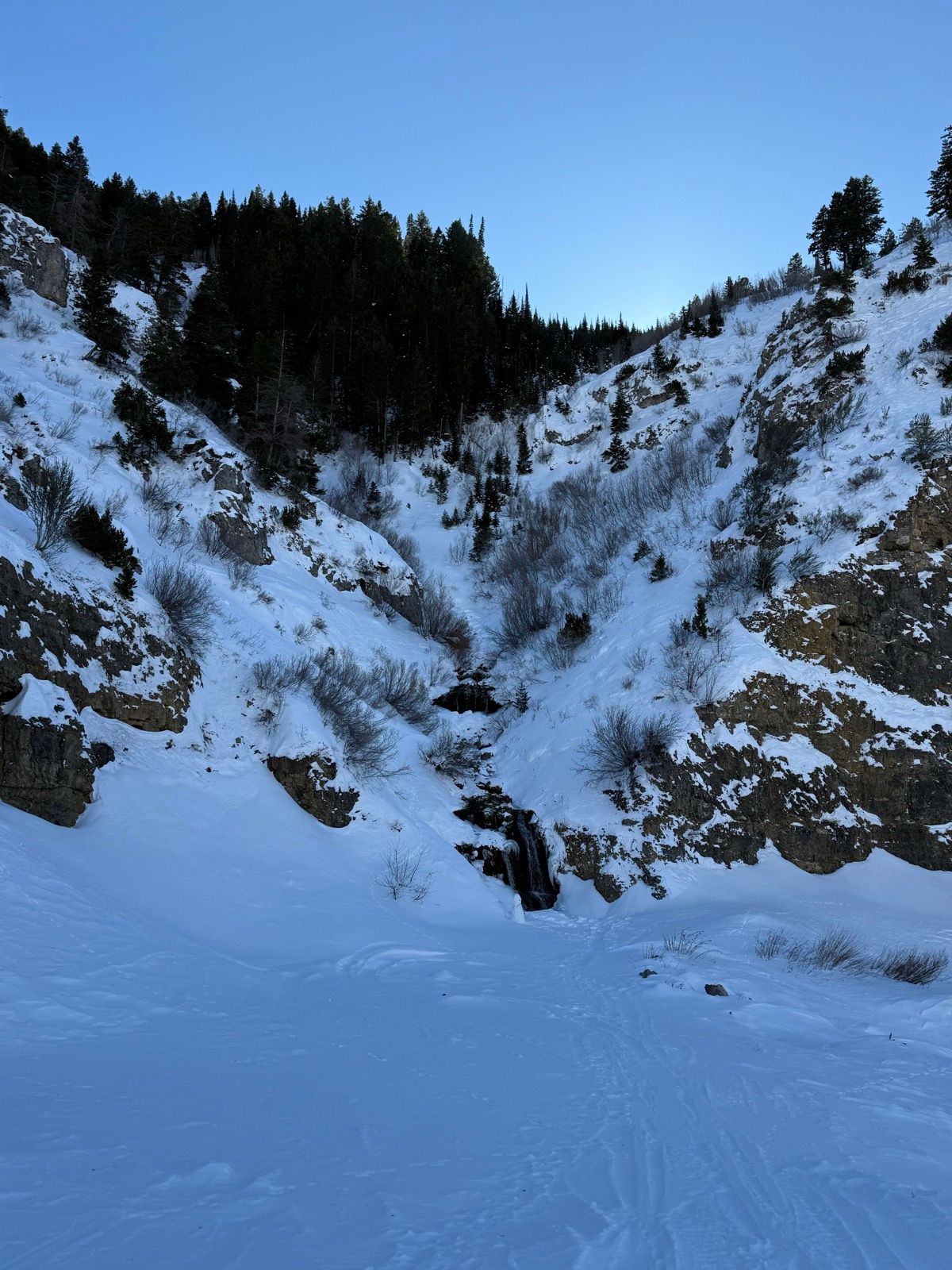 A snow-covered landscape in Providence, Utah, featuring rocky hills on either side. A small waterfall is visible in the center, flowing down the slope, surrounded by trees and shrubs. The sky is clear and blue.