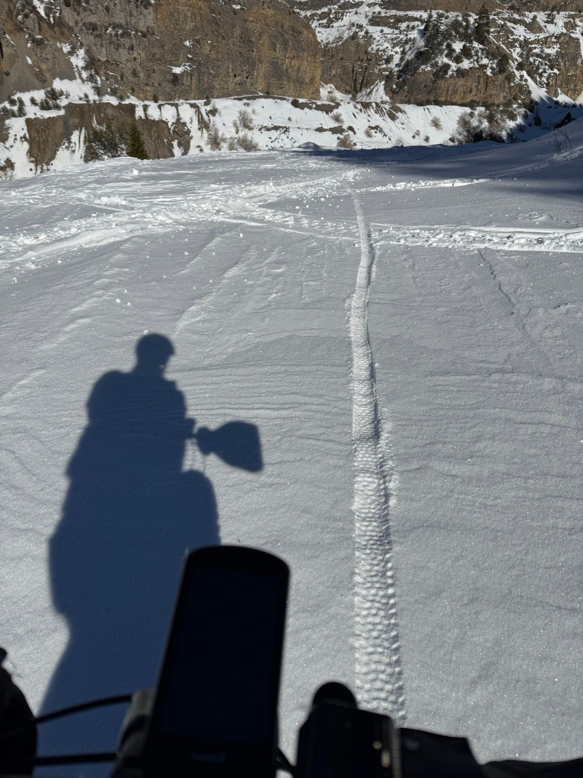 A snowy landscape in Providence, Utah, featuring tracks in the snow and a person's shadow. The foreground includes the handle of a snow shovel and the top of a snowmobile's dashboard.