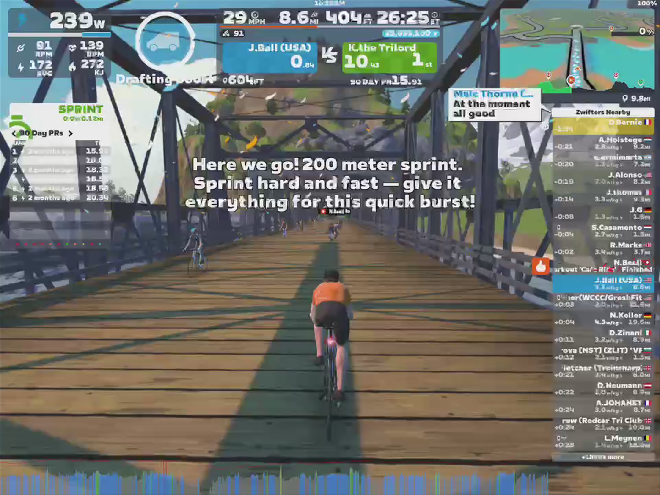 A cyclist in an orange shirt is participating in a virtual biking event on a wooden bridge within the Zwift app. The screen displays performance statistics, including power output and speed, along with a message encouraging a 200-meter sprint. Nearby cyclists are also visible in the scene.