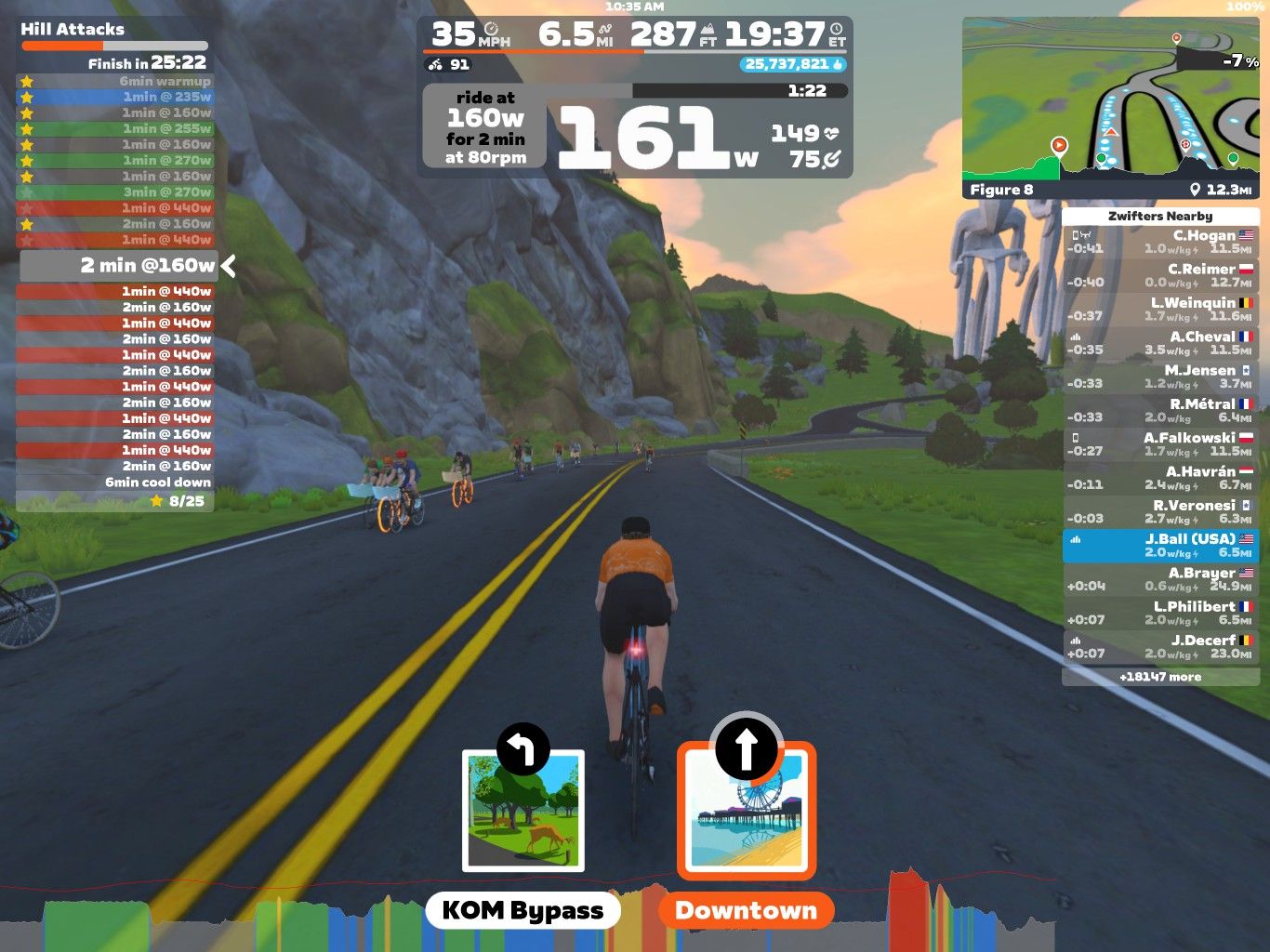 A cyclist in an orange shirt rides on a virtual road in Zwift, a cycling simulation platform. The screen displays performance metrics, including speed, distance, and wattage, along with a leaderboard on the right side showing other riders. Scenic graphics with rocks and trees are visible in the background.