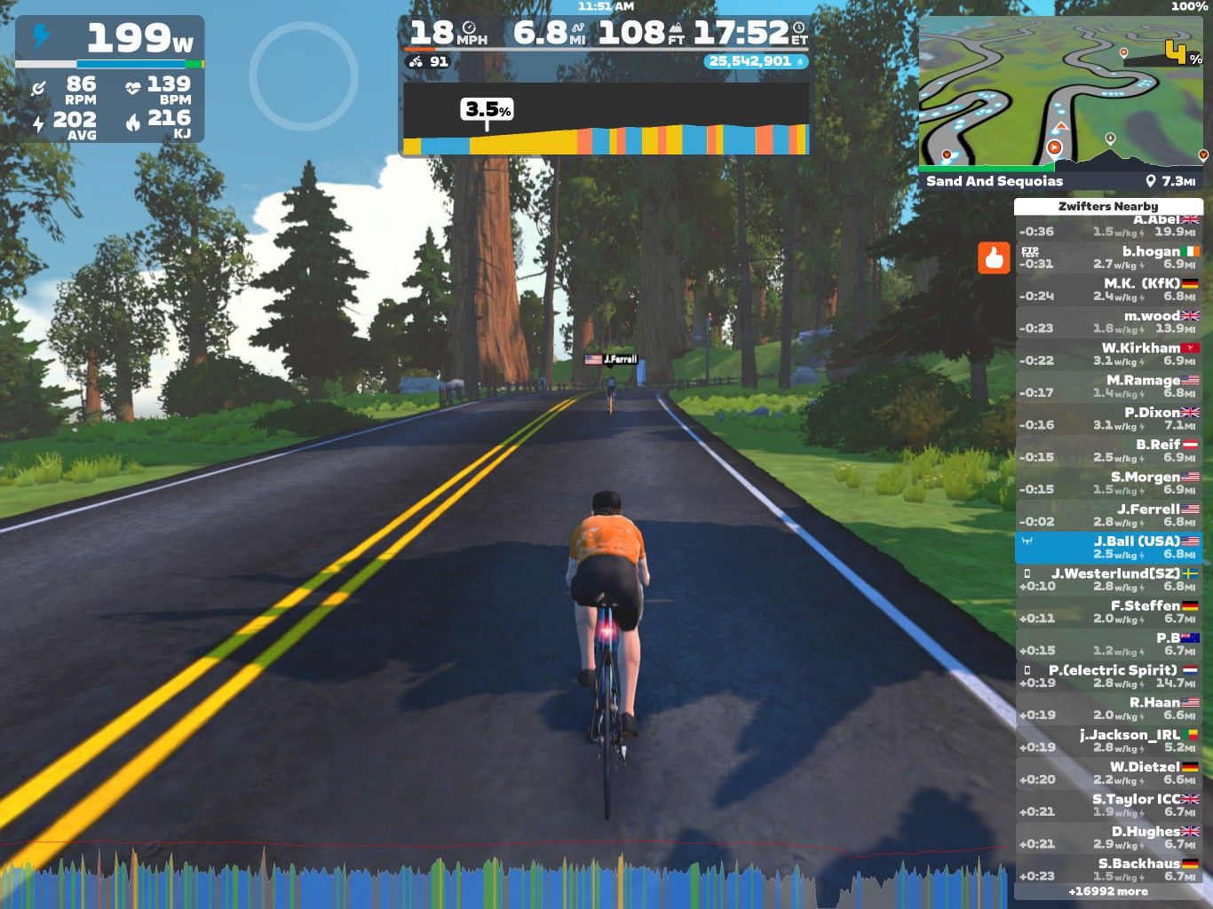 A cyclist wearing an orange shirt rides on a virtual road surrounded by trees in the Zwift app. The display shows various stats, including power output, speed, and distance traveled, along with a leaderboard of nearby riders. The scene captures a scenic, digital landscape intended for cycling workouts.