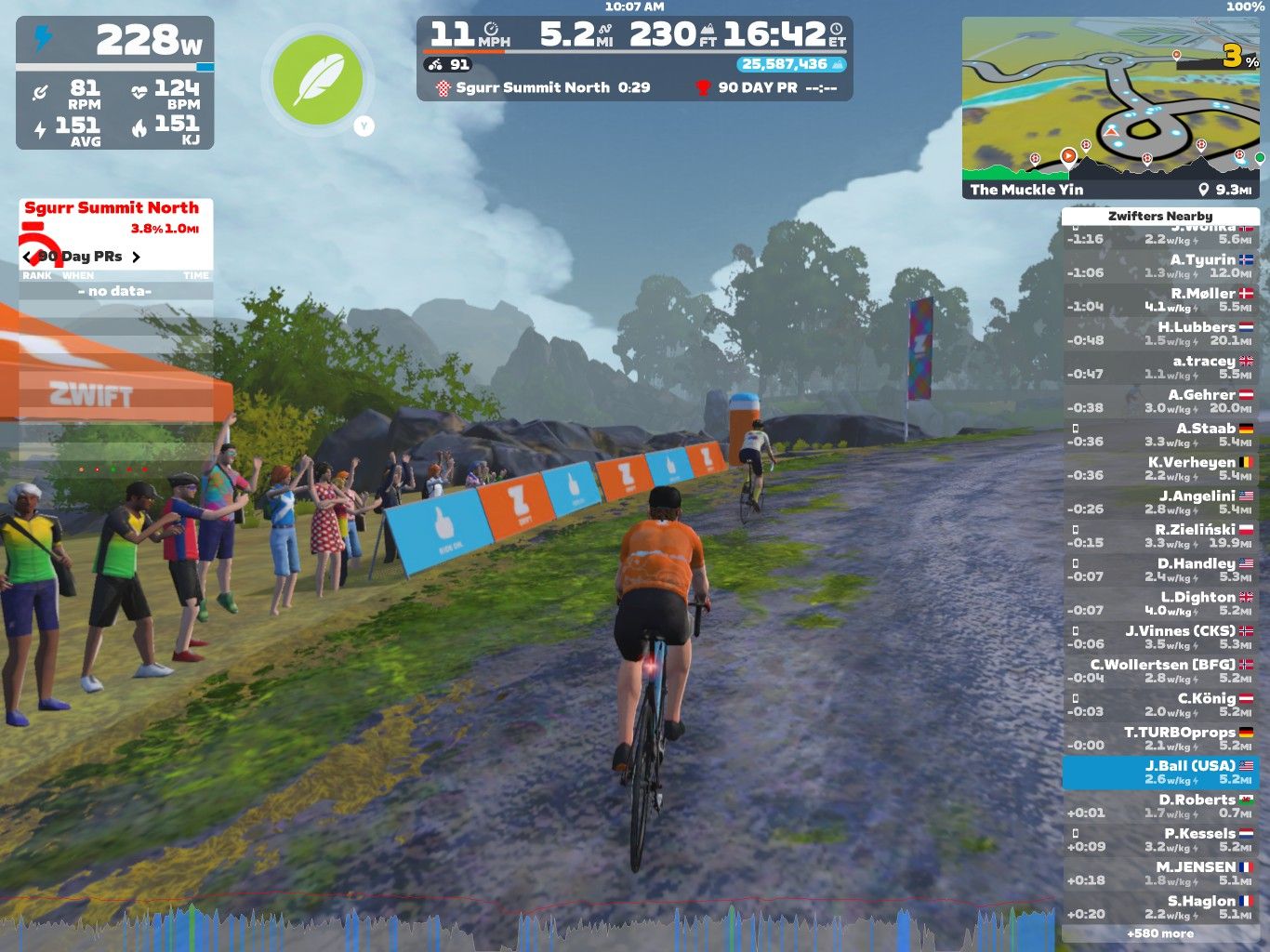 A cyclist in an orange shirt rides on a virtual dirt path in Zwift, surrounded by spectators cheering along the way. The scene includes a Zwift tent in the background, and a leaderboard showing nearby participants and their stats on the right side. The cyclists are moving through a lush, green landscape under a cloudy sky.