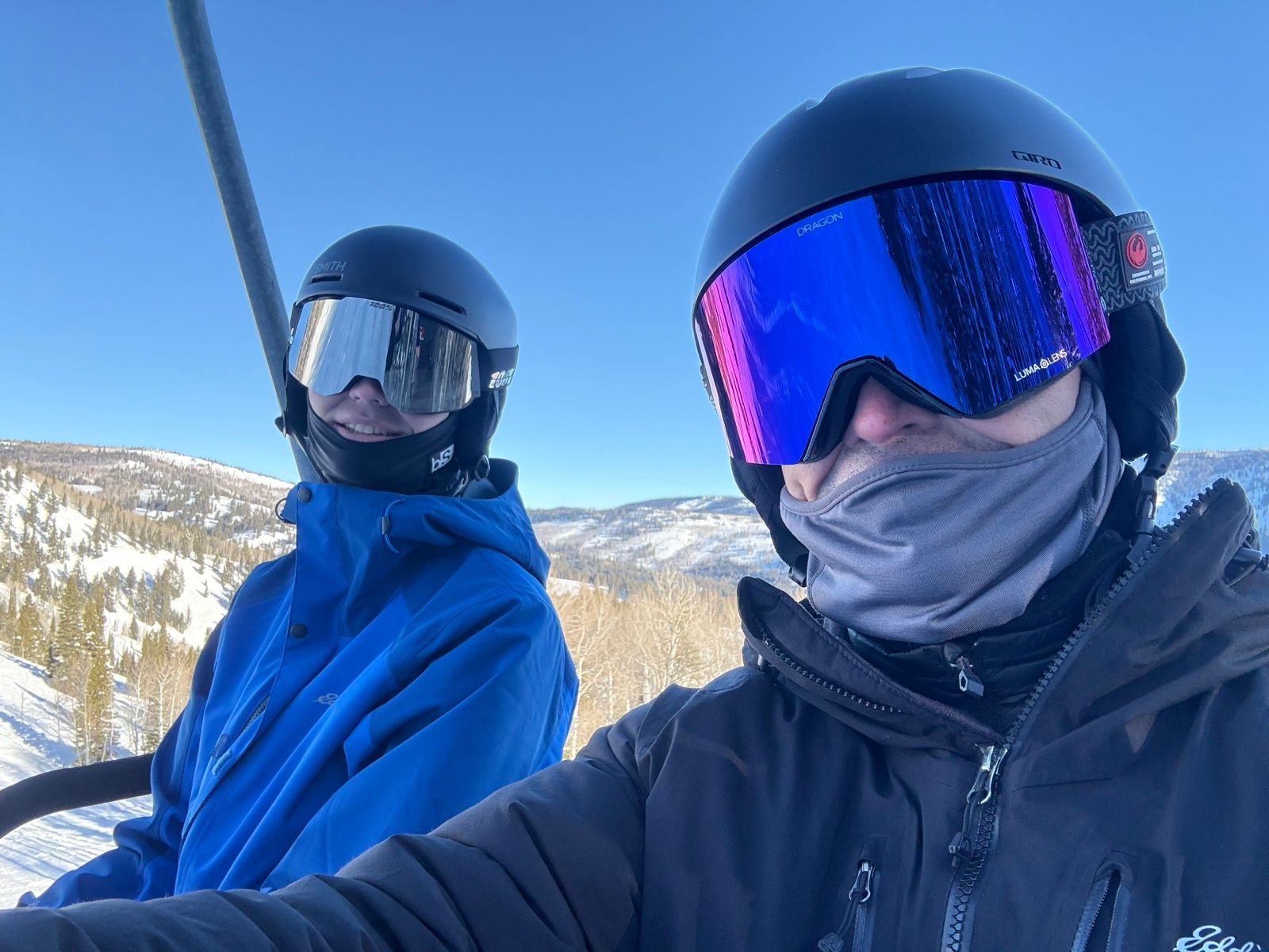 Two people are sitting on a ski lift, wearing helmets and goggles. One is in a blue jacket and the other in a black jacket, both smiling. Snow-covered mountains and clear blue sky are visible in the background.