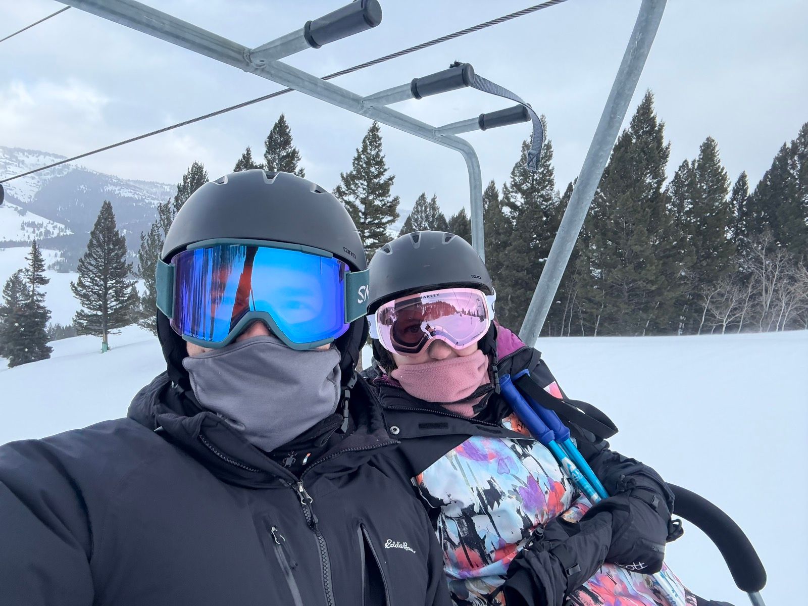 Two people wearing ski helmets and goggles ride a ski lift in snowy Logan, Utah. They are bundled up in winter clothing, with trees and mountains in the background.