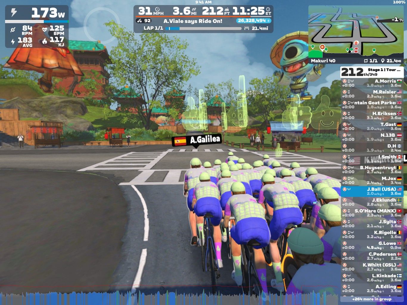 A group of cyclists in matching purple and green uniforms rides together on a virtual course in Zwift. The scene features colorful buildings and trees in the background, with a digital display showing race statistics, including speed, distance, and heart rate. The cyclists are navigating a turn, and there are virtual leaderboard details on the right side of the image.