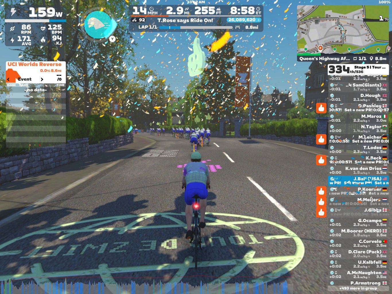 A cyclist is riding on a virtual road in Zwift, surrounded by other riders. The scene is bright and festive, with confetti falling from the sky. The interface shows performance metrics, including power output, heart rate, and speed, along with a leaderboard on the side. A map is visible in the upper corner, and the cyclist is approaching a turn on the course.