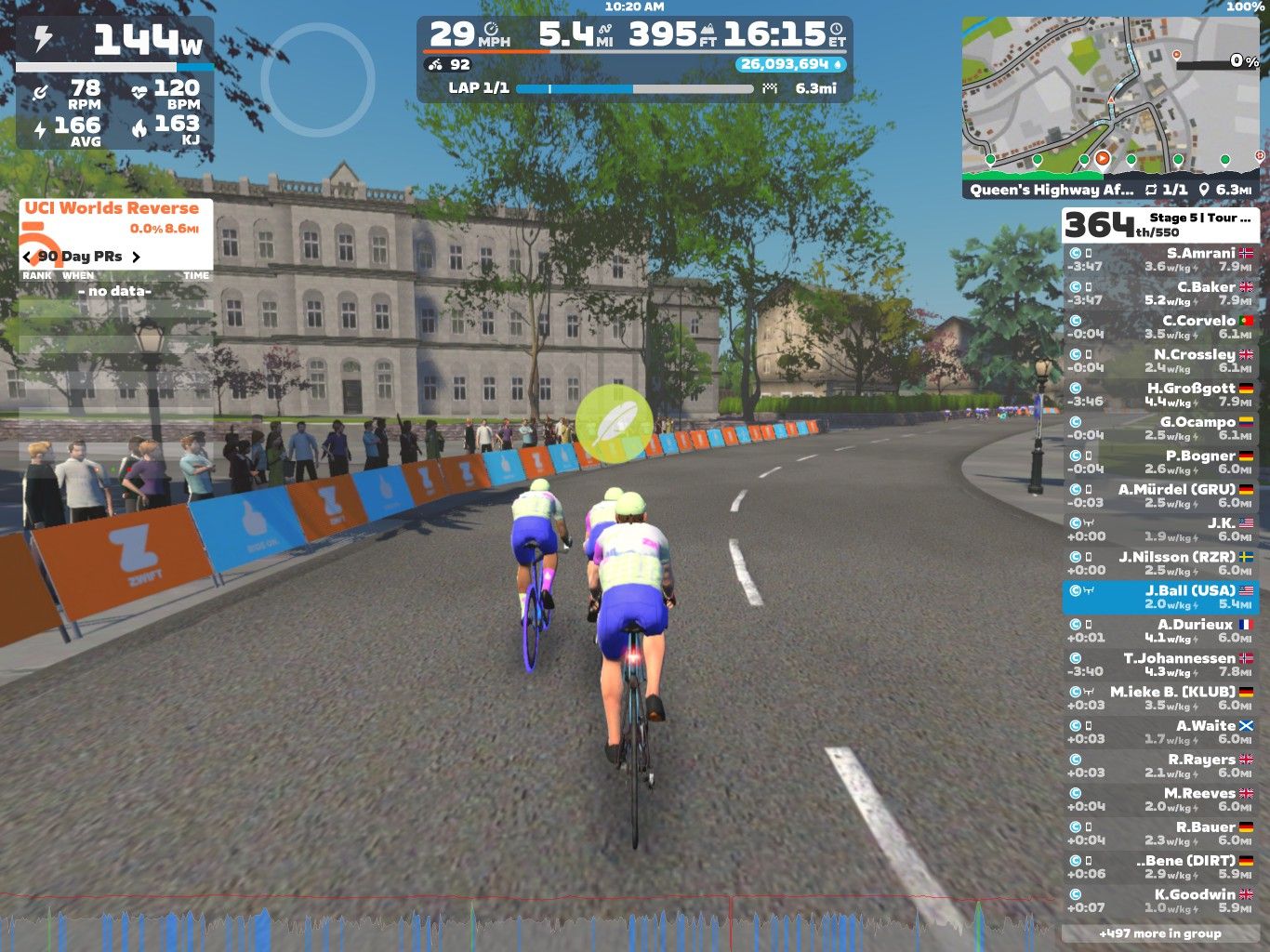 Two cyclists are riding along a virtual road in Zwift, surrounded by a crowd of spectators. The scene features a detailed backdrop of buildings and trees under a clear blue sky. The cyclists are wearing matching kits and are focused on their race, while various statistics are displayed on the screen, including speed, distance, and rankings. A map and additional race information are also visible in the corner.