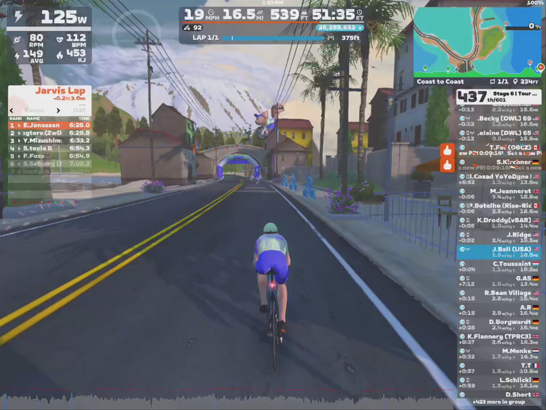 A cyclist rides on a virtual road in the Zwift application, with a visible leaderboard showing rankings and times. The scenery includes buildings, palm trees, and mountains in the background, with a blue sky above. The cyclist is wearing a green helmet and blue shorts, while statistics like power output and speed are displayed on the screen.