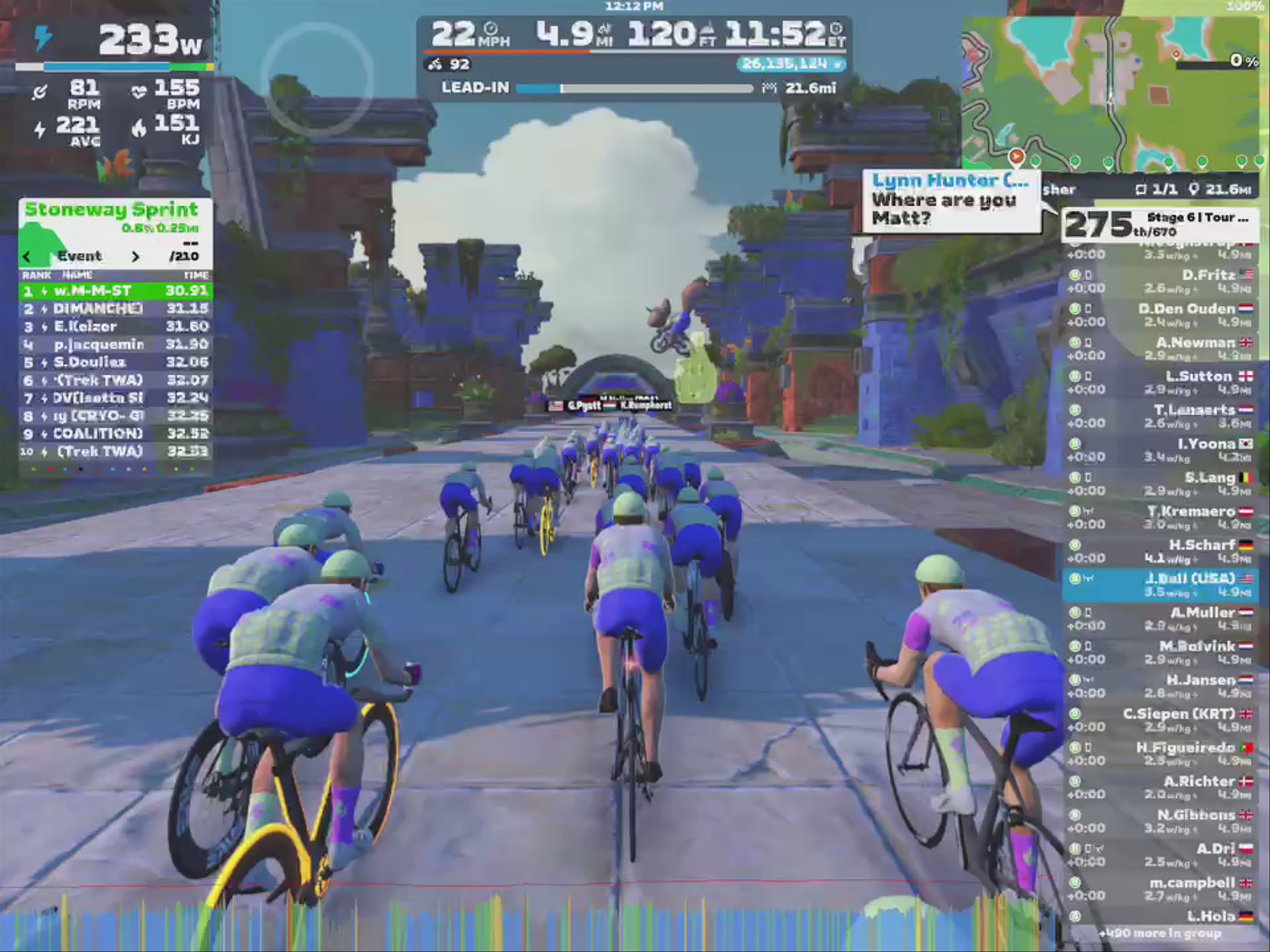 A digital cycling event on Zwift, showing a group of cyclists wearing blue jerseys riding on a virtual road surrounded by colorful, stylized buildings. The screen displays various statistics like power output, speed, distance, and a leaderboard of participants.