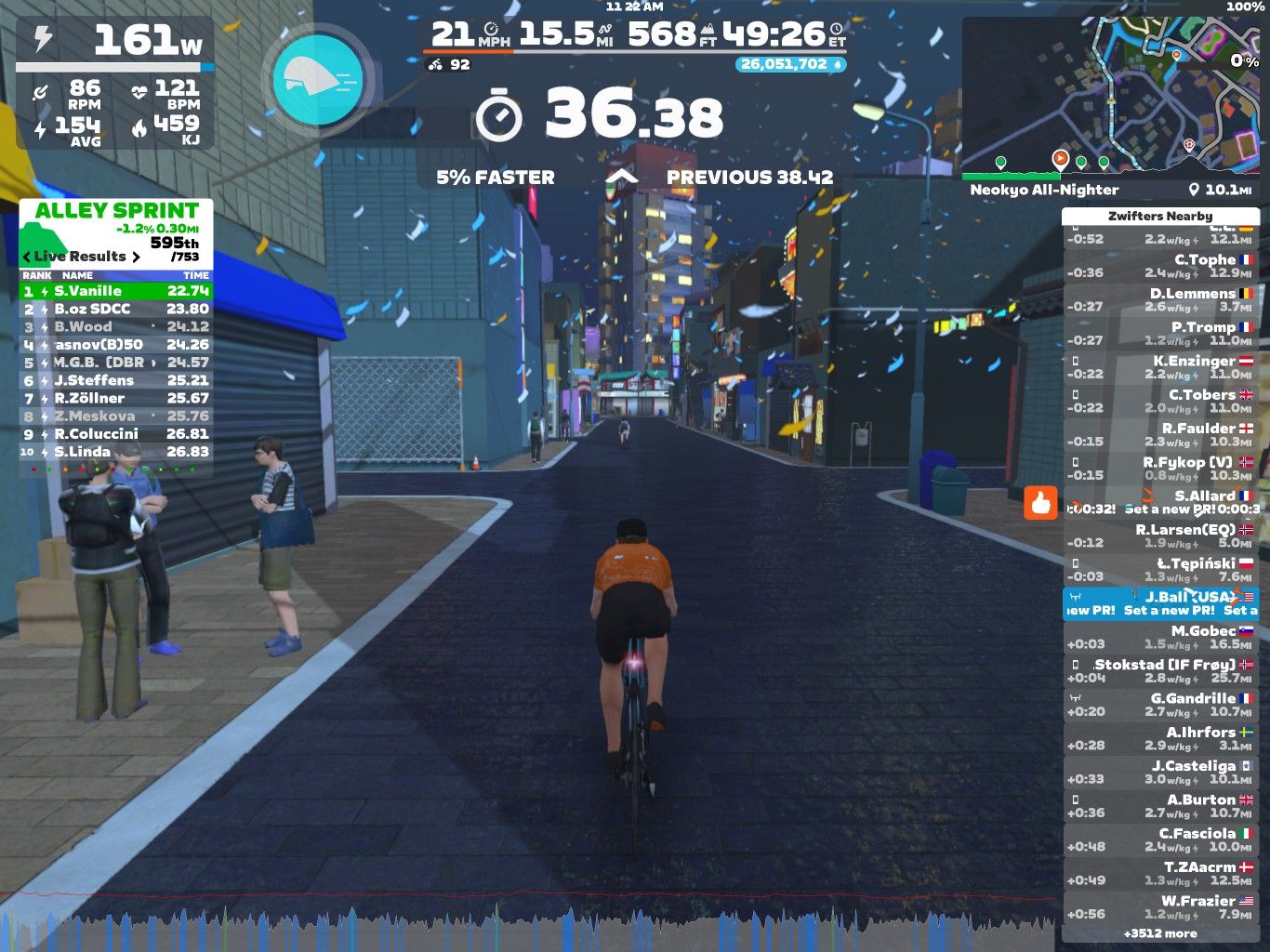 A cyclist in an orange jersey rides through a virtual urban street adorned with colorful buildings and confetti. The display shows speed, power, and heart rate data, alongside leaderboard results for a cycling event called