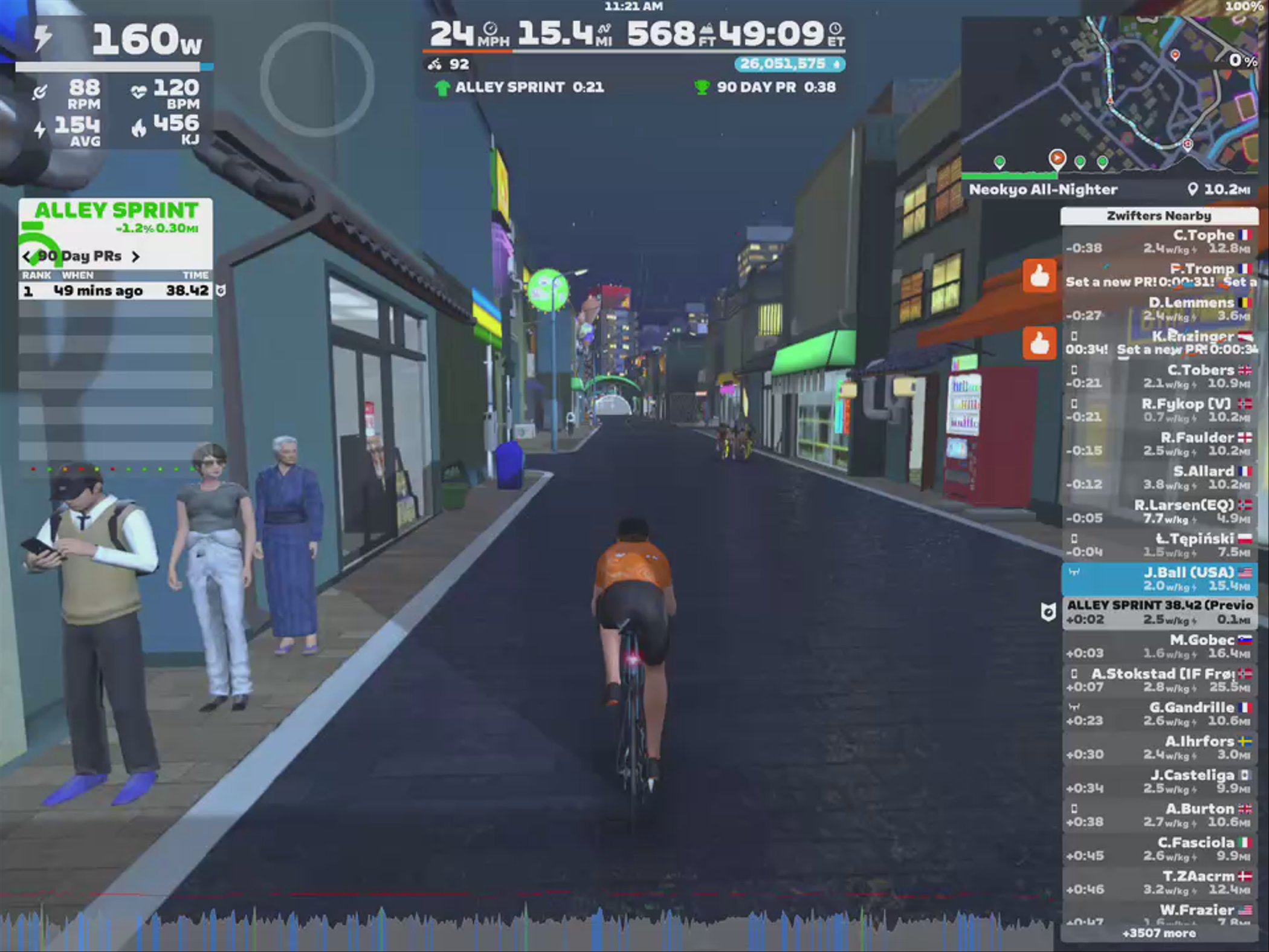 A cyclist in an orange jersey rides down a dark street in a virtual city, with various storefronts and colorful lights in the background. Pedestrians are visible on the sidewalk, with one person looking at a device. The scene displays cycling statistics and a map on the side, indicating an