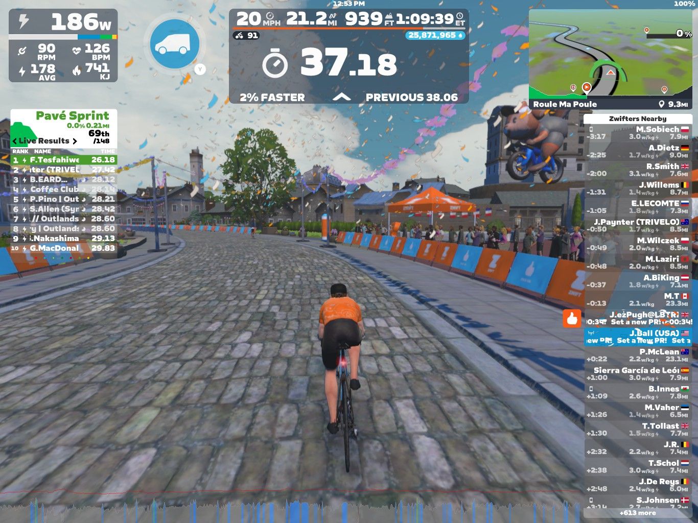 A cyclist in an orange jersey rides on a cobblestone path in a virtual race on Zwift. The scene includes colorful banners and spectators cheering, while digital stats display race progress, rankings, and nearby cyclists' performance. Confetti flutters in the air as the cyclist approaches a sprint segment. A map is shown in the corner, indicating the cyclist's location on the course.