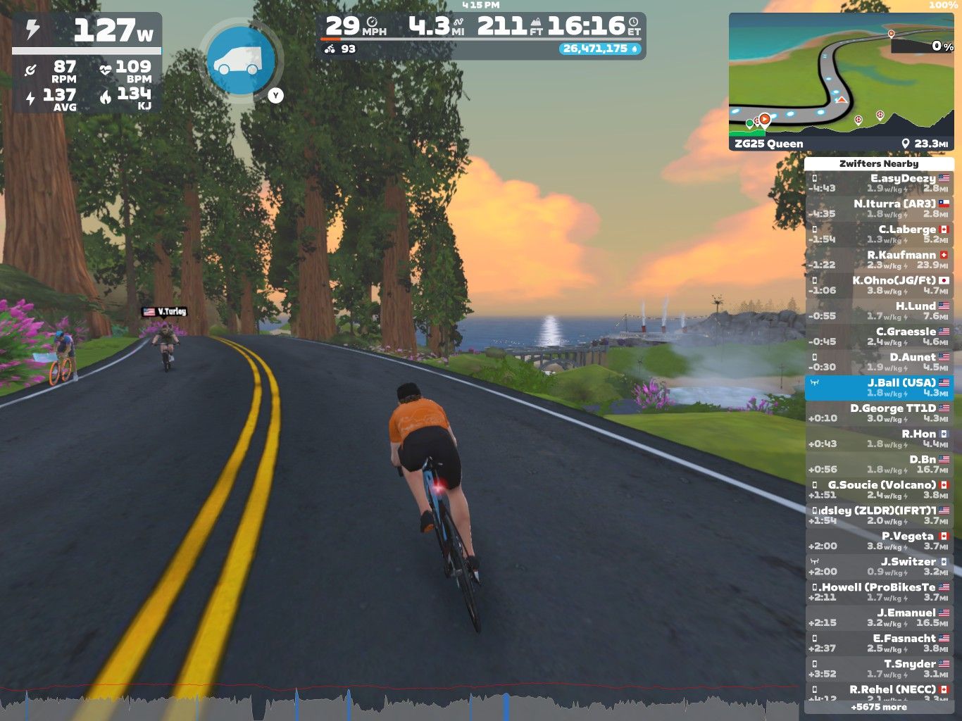 A virtual biking scene in Zwift featuring two cyclists on a winding road surrounded by trees and flowers. The display shows performance metrics, including power output, speed, and heart rate, alongside a map of the cycling route and a list of nearby participants. The sunset creates a colorful sky over the landscape.