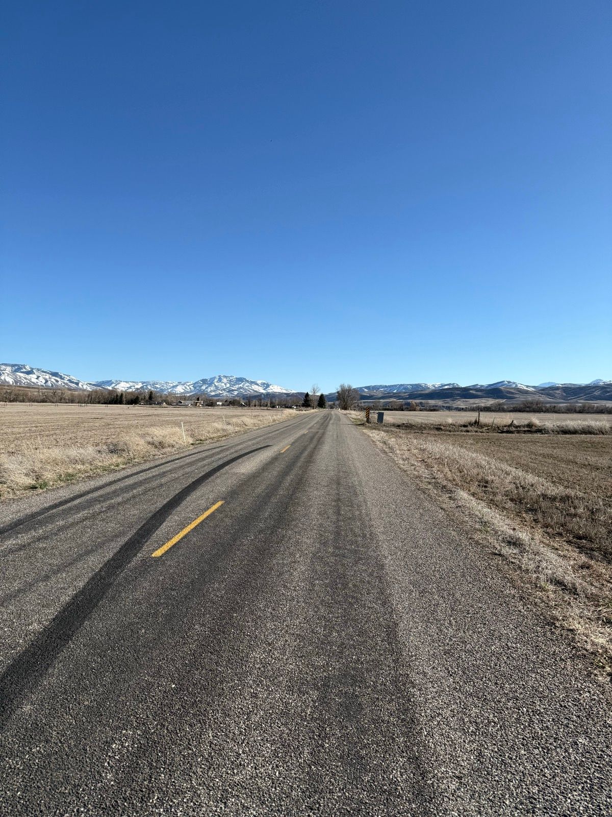 A straight, empty road stretches into the distance under a clear blue sky, surrounded by fields and mountains in the background. The landscape is mostly dry, with some patches of snow visible on the mountains.