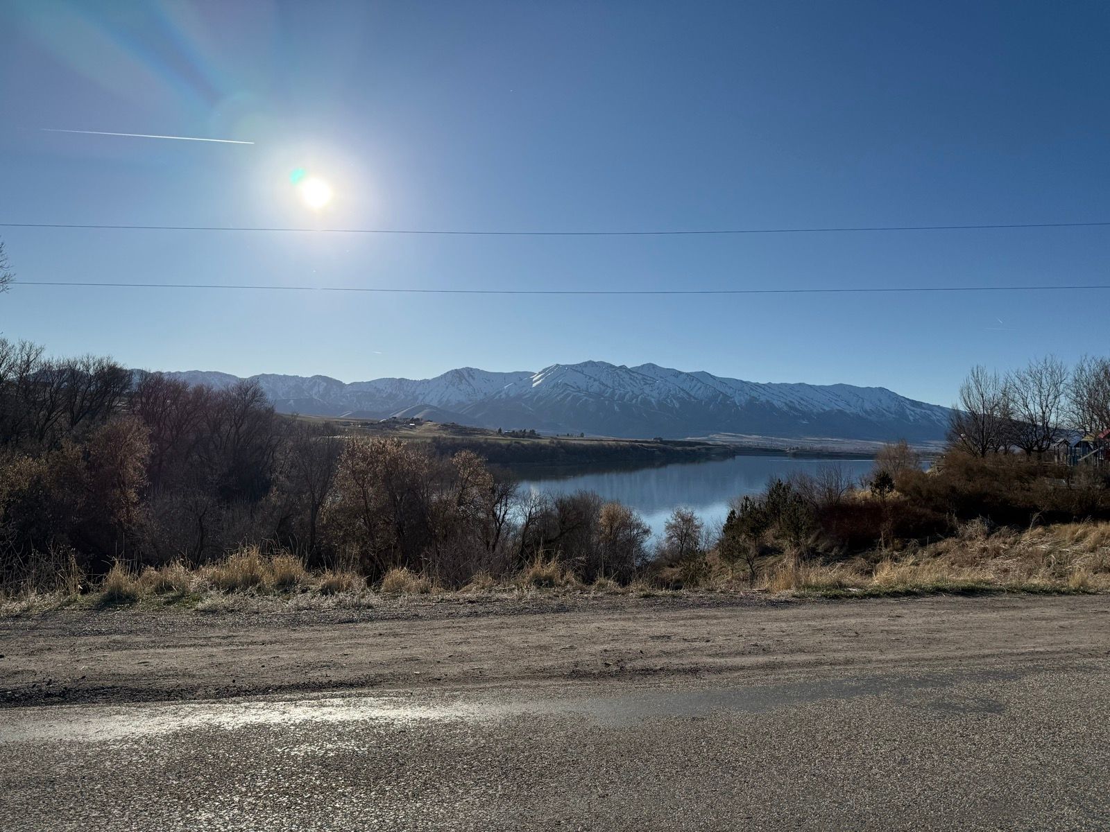 A tranquil view of a lake surrounded by mountains in Nibley, UT, under a clear blue sky. The sun is shining brightly, reflecting off the water, with patches of dry grass and trees in the foreground.