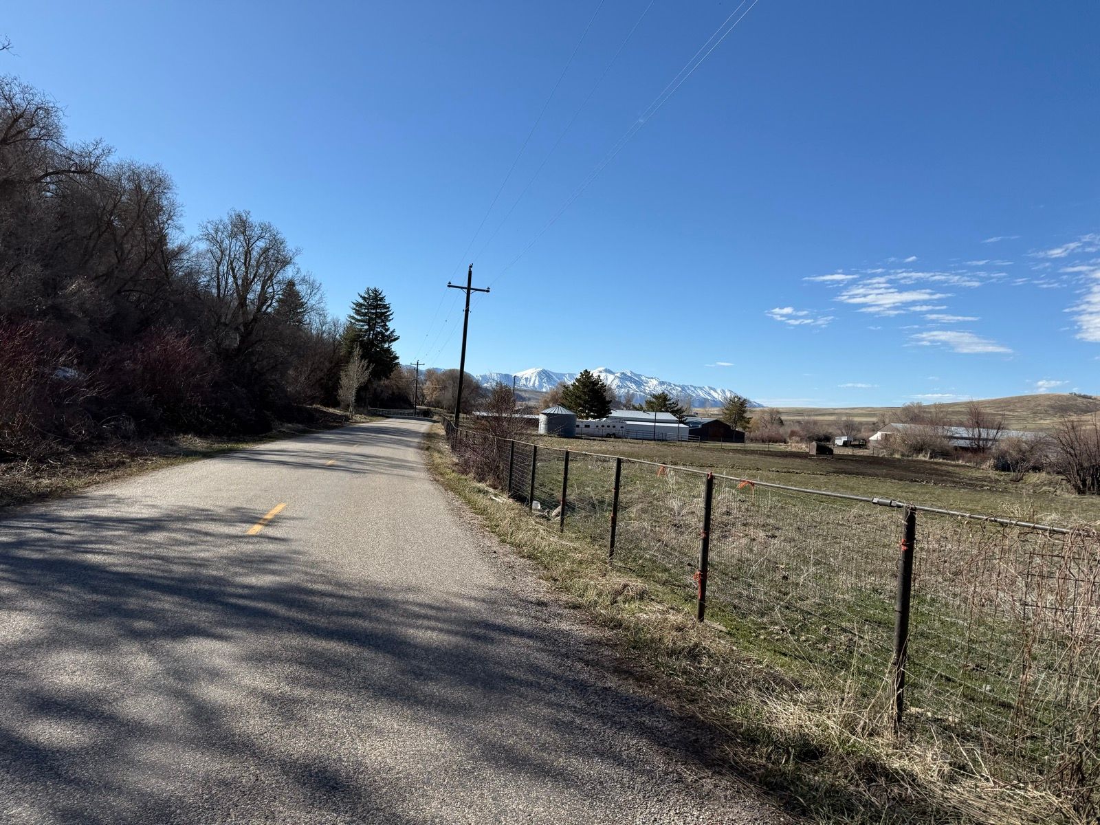 A quiet rural road stretches alongside a fence with sparse vegetation and trees lining one side. In the distance, fields and farm buildings are visible against a backdrop of snow-capped mountains under a clear blue sky.