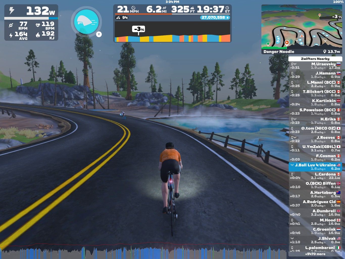 A cyclist in an orange jersey rides along a winding road in a virtual landscape with trees and a body of water. The screen displays various performance metrics, including speed, power, and heart rate, as well as a mini-map and nearby cyclists. The environment has a cloudy sky and rocky terrain.