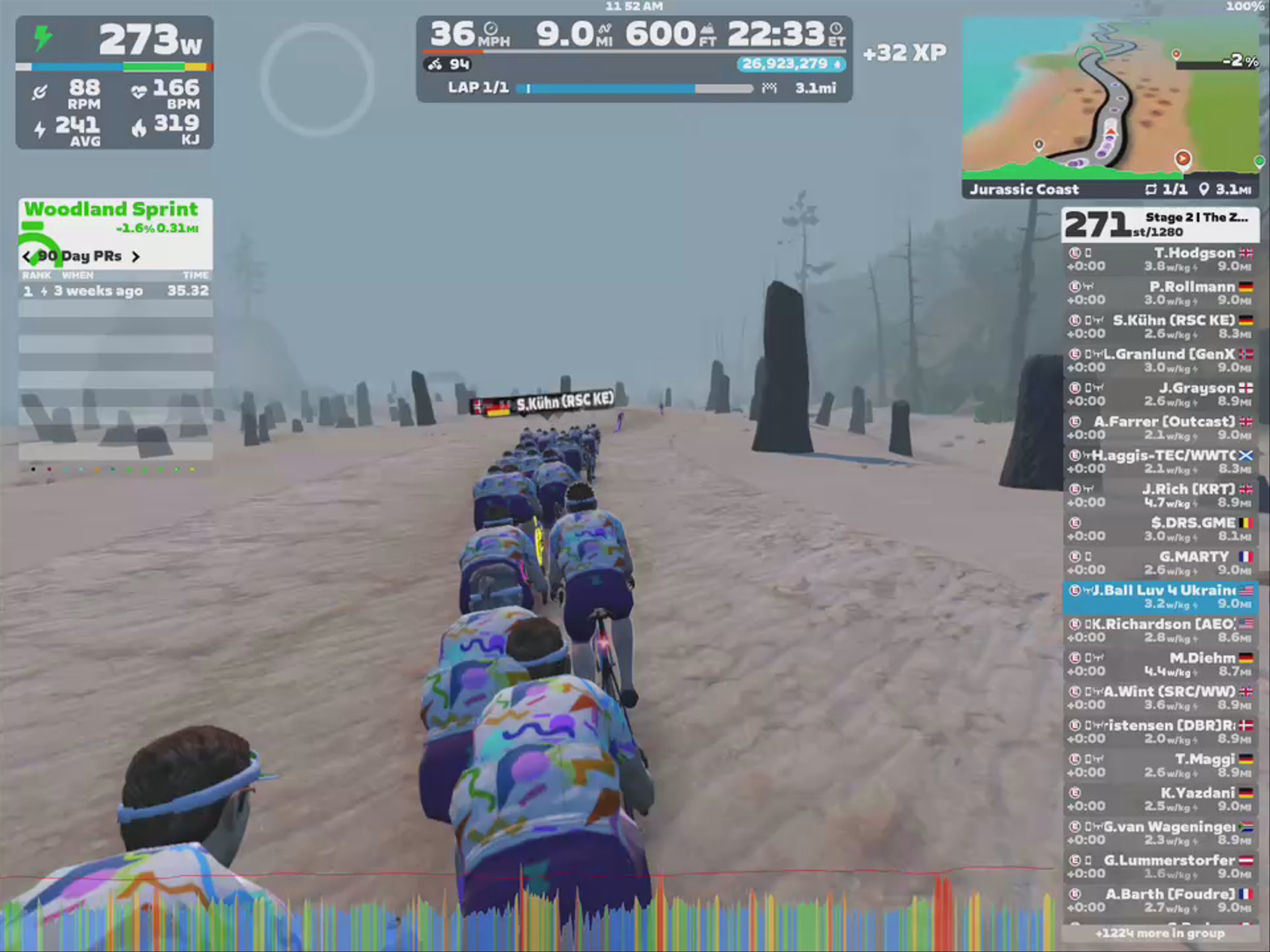 A group of cyclists in colorful jerseys is riding in a virtual landscape on Zwift, surrounded by stone formations and a foggy atmosphere. The screen displays cycling stats such as power, speed, and distance, along with a mini-map of the route.