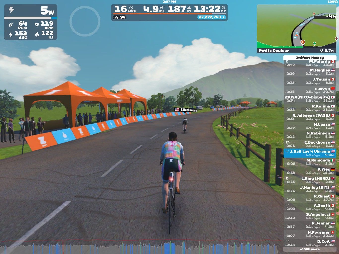A virtual cycling scene in Zwift, showing two cyclists on a winding road. The path is lined with cheering spectators and orange tents. The sky is blue with some clouds, and a mountainous background can be seen. Performance data and nearby cyclists' information are displayed on the right side of the image.