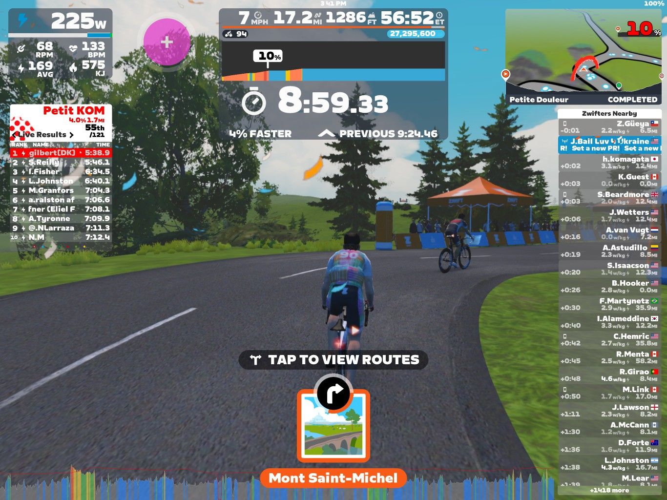 A digital cycling scene from Zwift shows riders pedaling on a virtual road with a leaderboard and various statistics displayed. The current time is 8:59.33, and the riders are near the location named Petite Douleur in the Zwift environment. There's a blue and orange tent on the side of the road, with trees and greenery in the background.