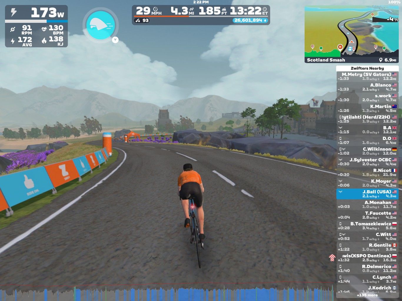 A cyclist in an orange jersey rides on a winding virtual road in Zwift, with mountains and trees in the background. The display shows cycling statistics and nearby riders. Colorful banners line the road, promoting the event.