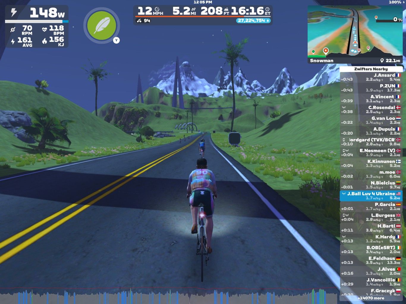 A cyclist is riding on a virtual road in Zwift, surrounded by green hills and mountains. The screen displays speed, power output, and other performance metrics. Nearby cyclists are listed on the right side. The setting is colorful with a clear blue sky and palm trees, indicative of a scenic environment.