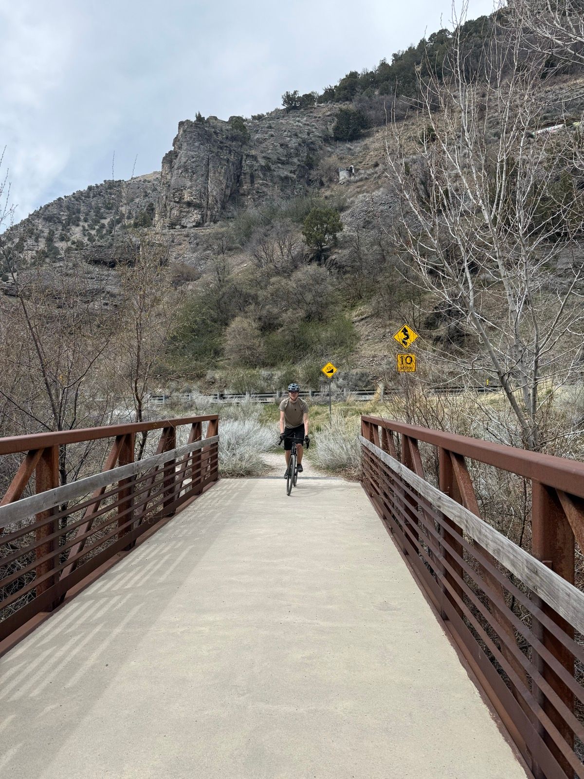 A cyclist rides across a metal bridge in Paradise, Utah, surrounded by rocky hills and sparse vegetation. The sky is overcast, and there are roadside warning signs in the background.