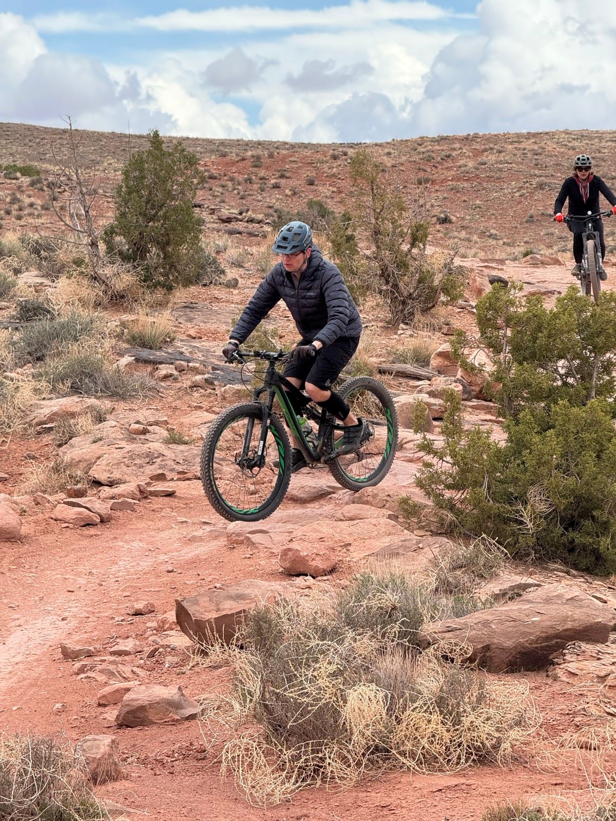 Two mountain bikers ride on a rocky trail in Moab, Utah. The first rider, wearing a blue helmet and dark clothing, is navigating down a slope, while the second rider, further back, is also on the trail. The landscape features reddish dirt, sparse vegetation, and a cloudy sky.