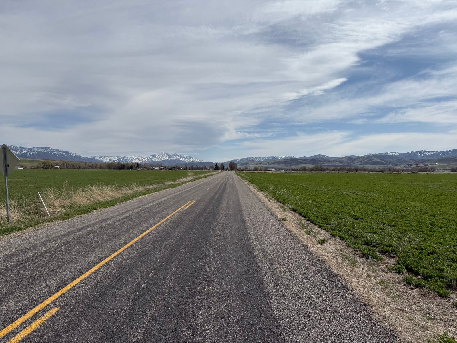 A rural road stretches through green fields in Nibley, Utah, with mountains in the background and a cloudy sky overhead.