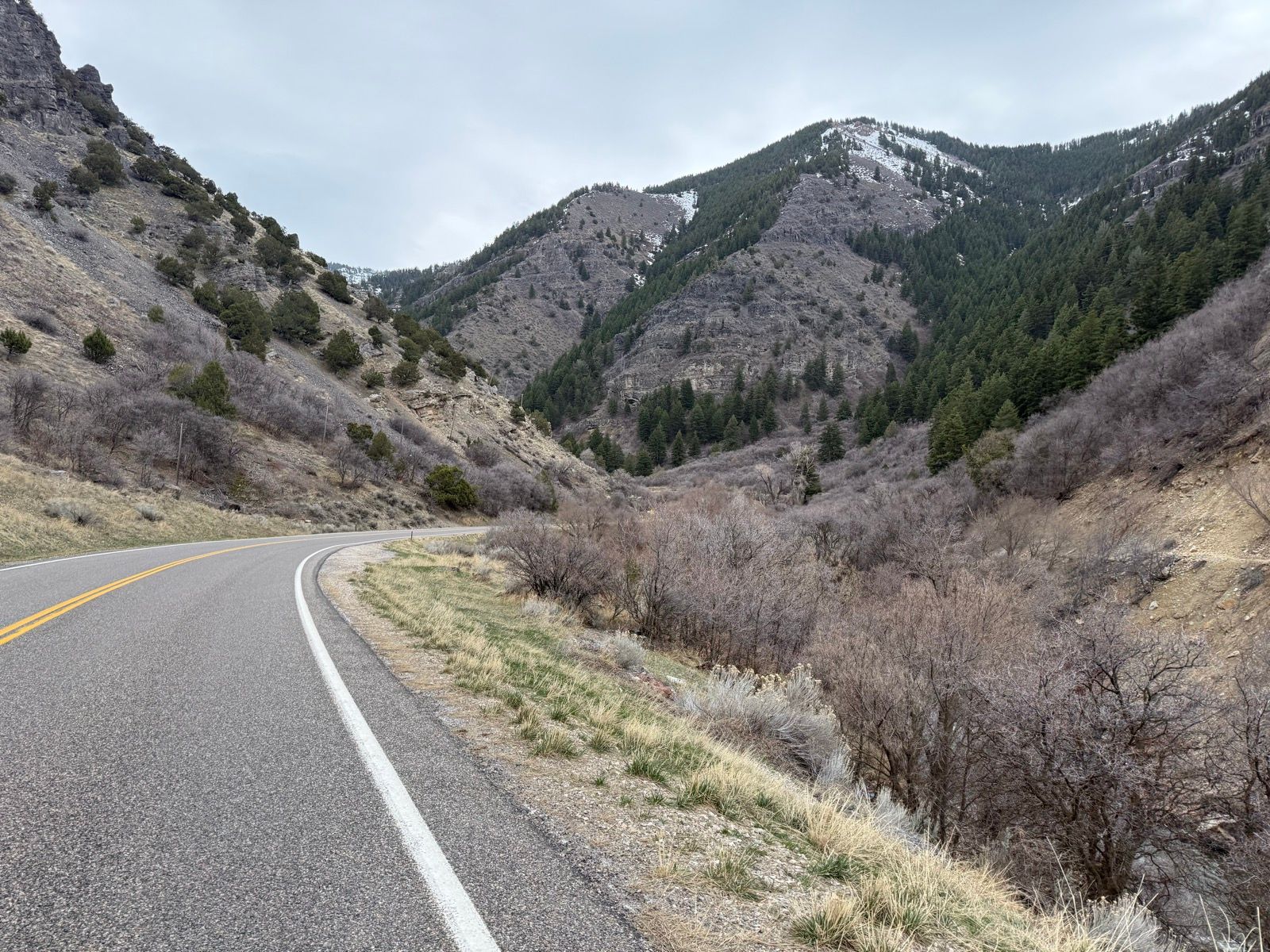 A winding road surrounded by mountains in Nibley, Utah. The landscape features rocky hills with sparse vegetation and some evergreen trees, under a cloudy sky.