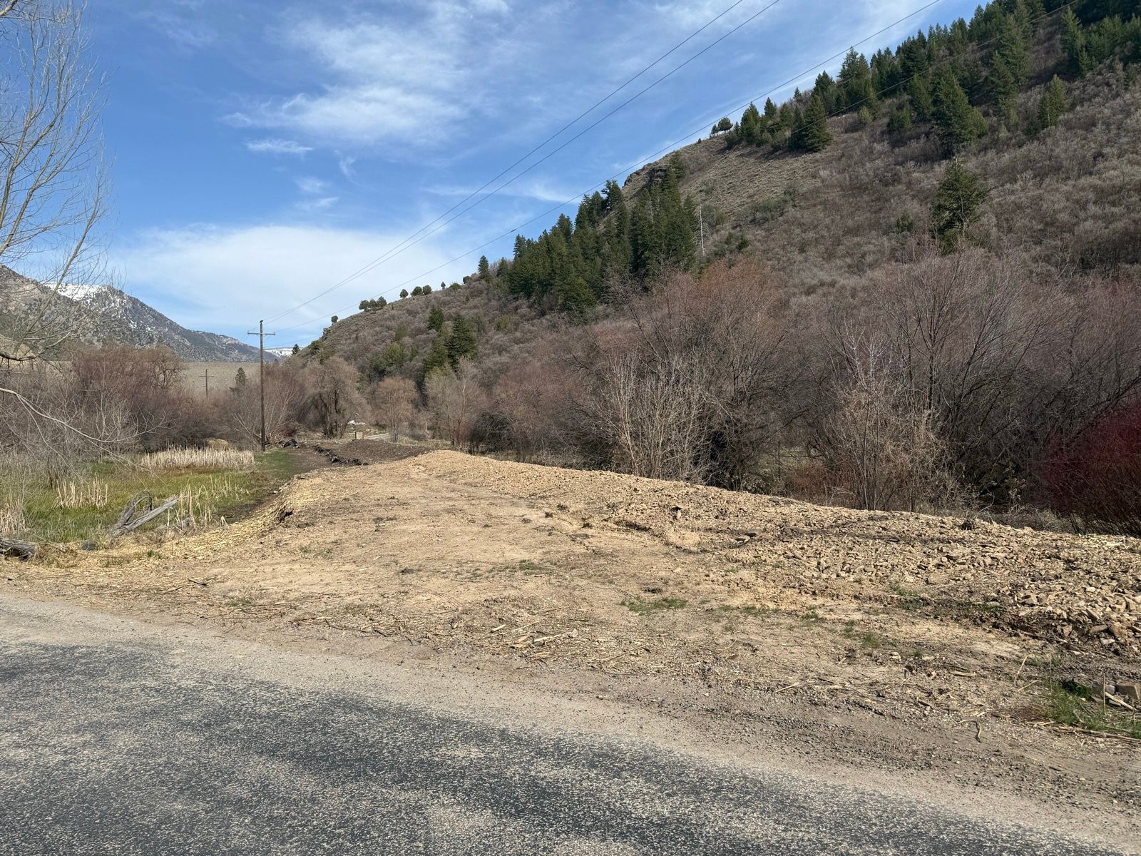 A rural landscape in Nibley, Utah, featuring a gravelly area with sparse vegetation, bordered by trees on both sides. In the background, there are rolling hills and distant snow-capped mountains under a partly cloudy sky. Utility poles are visible along the road.