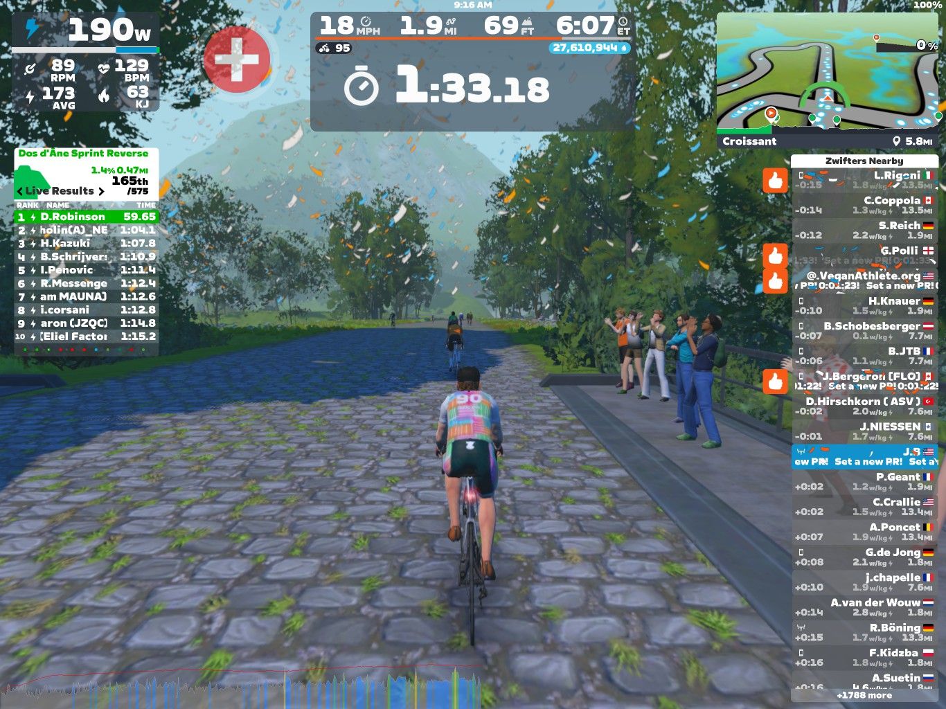 A cyclist rides on a virtual cobblestone path in Zwift, surrounded by green trees and digital spectators. The screen displays cycling stats, such as power output, speed, and heart rate, along with a leaderboard and a map of the area.