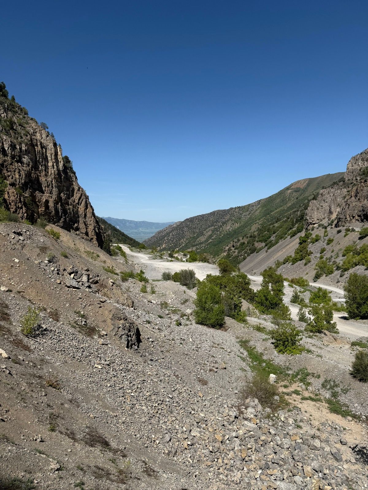 A view of Providence Canyon in Utah, showcasing rocky terrain, steep hillsides, and a clear blue sky. Sparse vegetation is visible on the canyon slopes, and the canyon floor appears dry and gravelly.