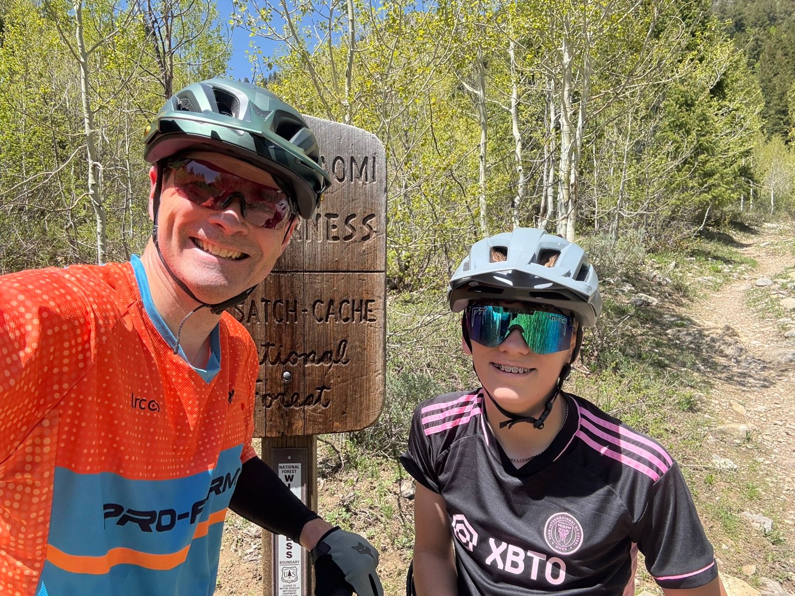 A man and a young girl wearing helmets and sunglasses pose for a selfie at a trail sign in Green Canyon, Utah. The sign indicates the area is part of the Wasatch-Cache National Forest, surrounded by lush greenery.