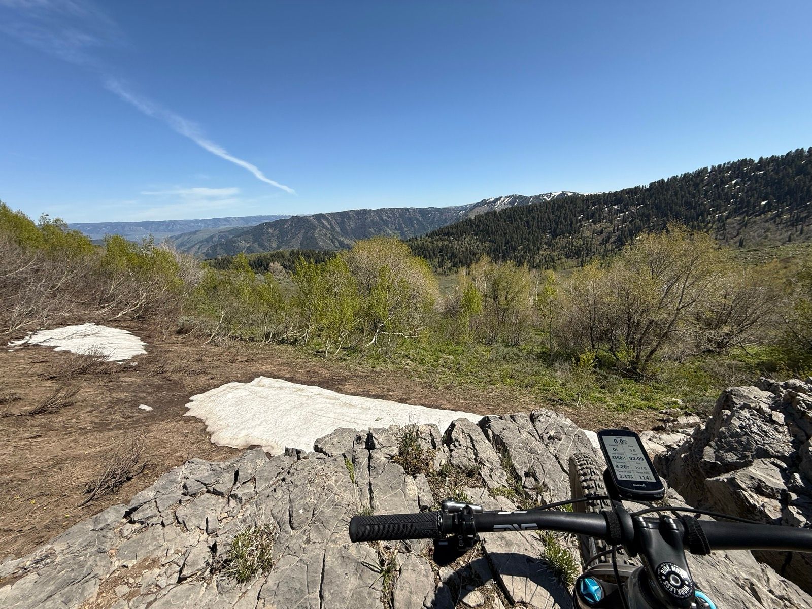 A scenic view from a rocky overlook in Providence Canyon, Utah, featuring distant mountains, green vegetation, and patches of remaining snow on the ground. A mountain bike is positioned in the foreground, displaying a device with biking stats. The sky is clear with a few wispy clouds.