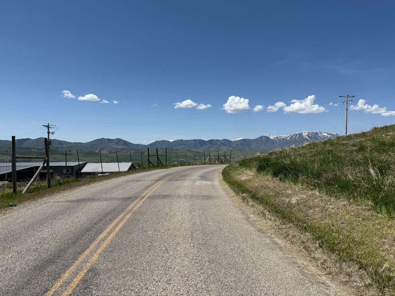 A winding road curves through a rural landscape in Nibley, Utah, with fields on either side and mountains in the background. The sky is clear with a few scattered clouds. Utility poles line the road, and a fence runs alongside the fields.