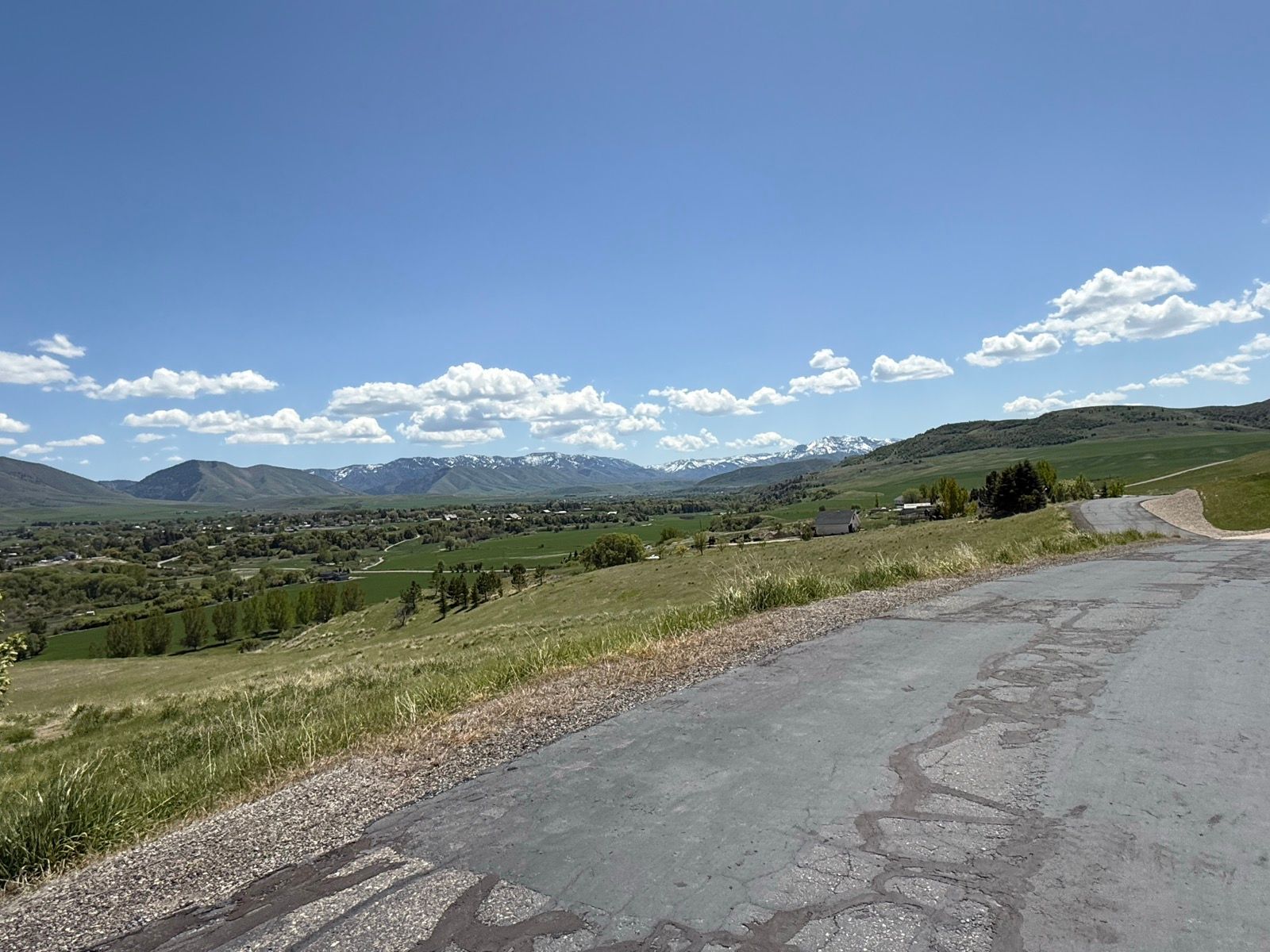 A scenic view of a valley in Nibley, Utah, featuring green fields, distant mountains with snow, and a clear blue sky with fluffy clouds. A winding road leads through the foreground, surrounded by grassy areas and scattered trees.