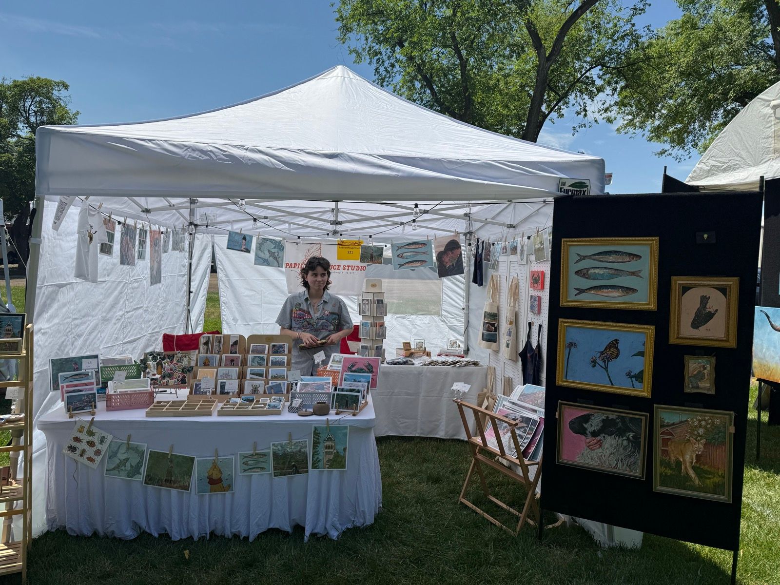A vendor stands behind a table at an outdoor art fair under a white tent in Providence, Utah. The table is covered with various art prints and greeting cards. In the background, an art display featuring framed pieces hangs on a black panel. The sky is blue with a few clouds, and there are trees in the background.