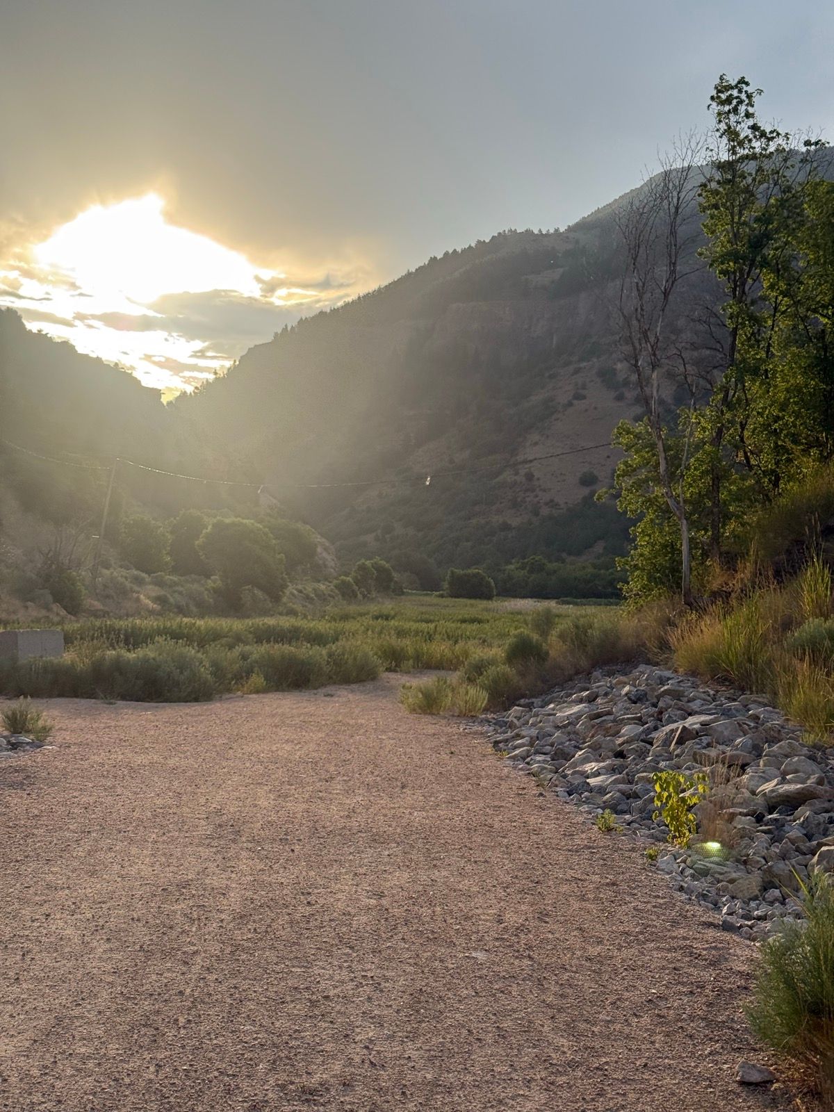 A gravel path in Paradise, Utah, leads towards a view of mountains under a cloudy sky, with the sun setting behind the peaks. Lush greenery and a rocky area are visible on either side of the path.