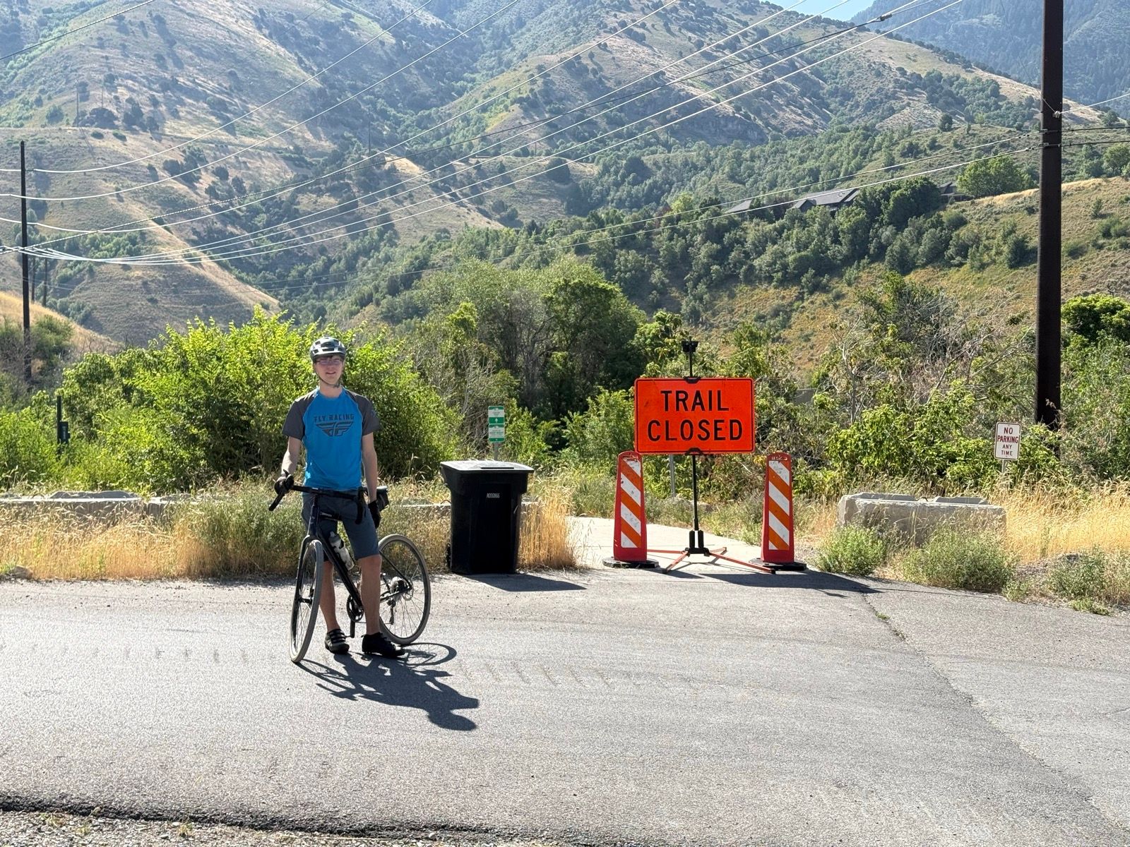 A person in a blue t-shirt and helmet stands next to a bicycle at a road intersection. There is a bright orange 