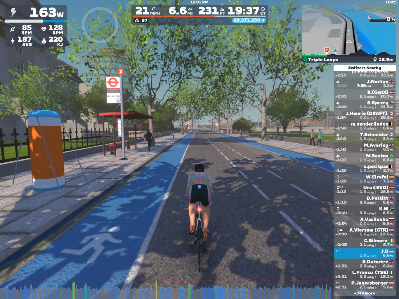 A cyclist rides along a blue bike lane in a virtual environment, with trees lining the road. There is a bus stop nearby, and a few pedestrians are visible in the background. The cyclist is wearing a colorful jersey, and a statistics panel shows performance data such as speed and power.