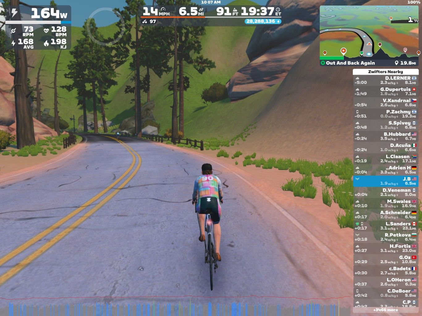 A cyclist rides on a winding road surrounded by green hills and trees in a virtual environment, displaying cycling statistics on the screen. The interface shows speed, heart rate, and other performance metrics.