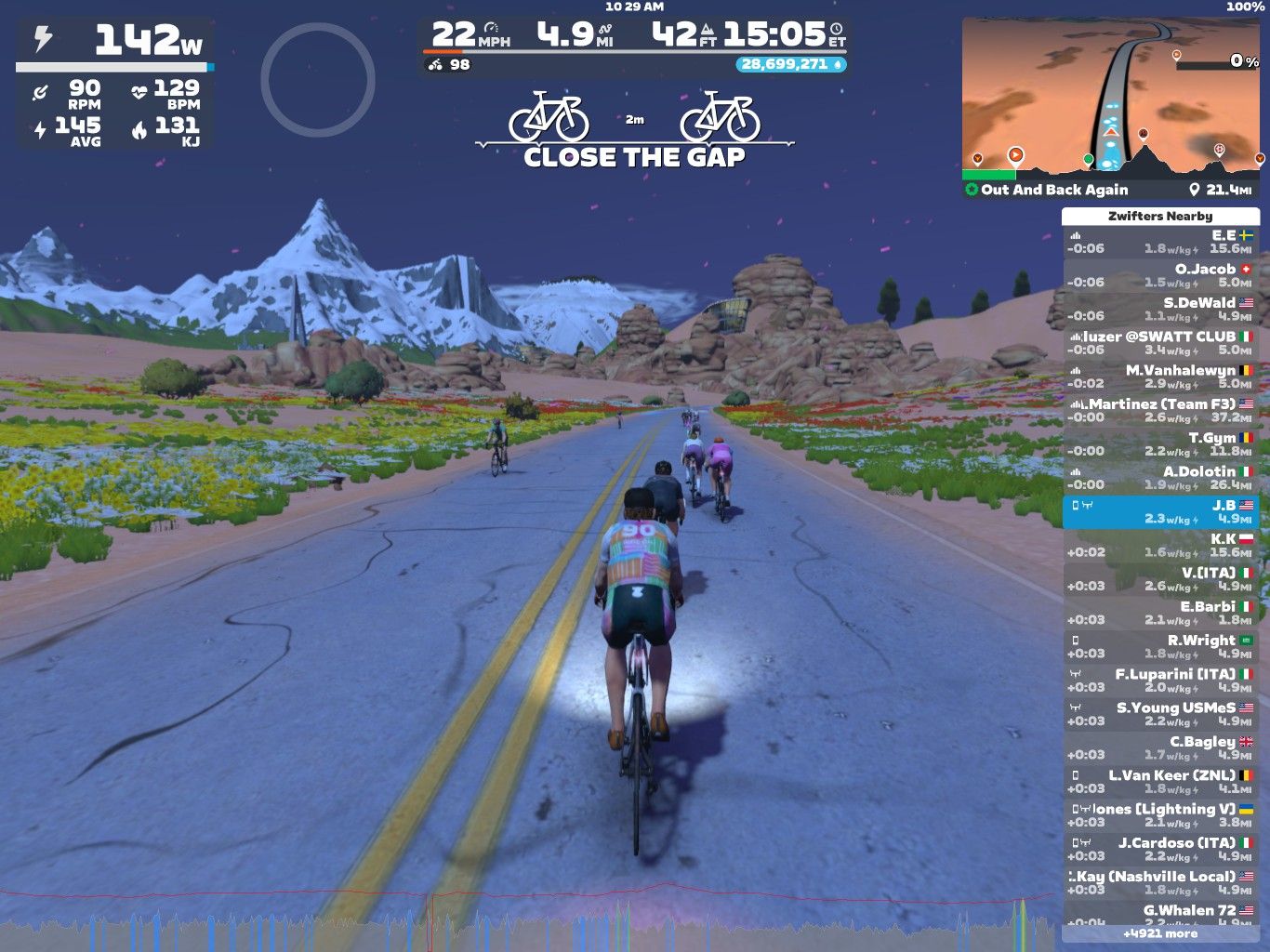 A group of virtual cyclists rides along a smooth, winding road surrounded by colorful flowers and grassy fields, with snow-capped mountains in the background. The scene is set in Zwift, featuring digital stats like speed, distance, and power output on the screen.